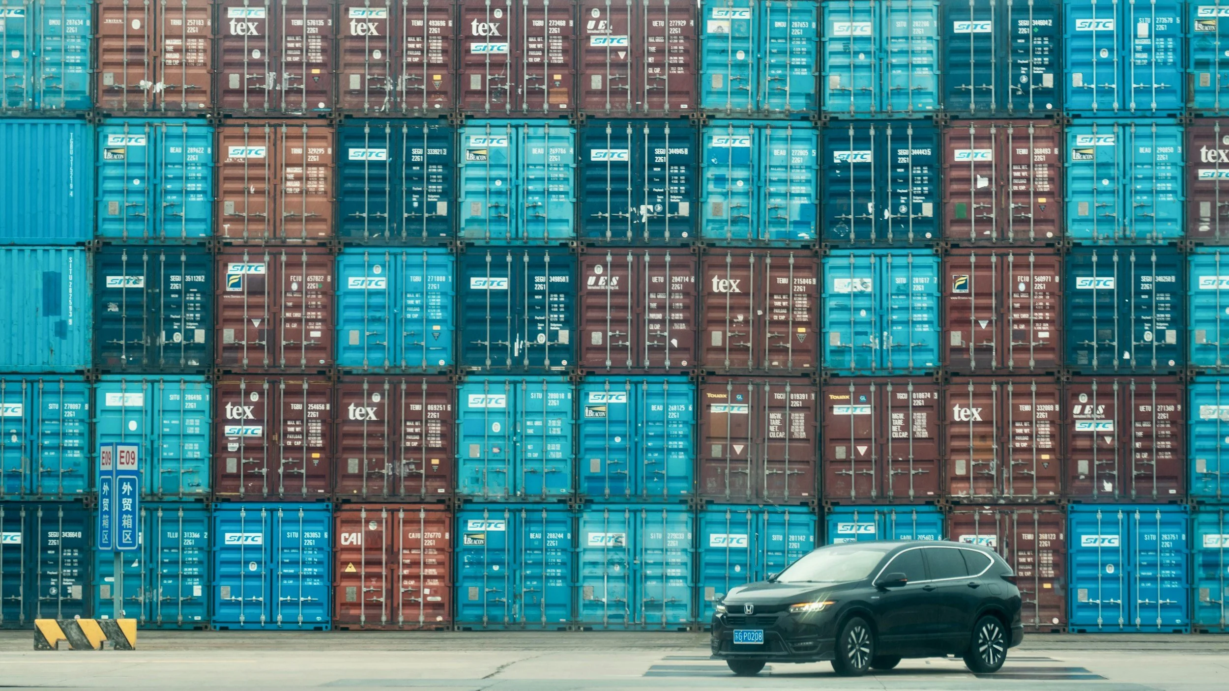 A black SUV parked in front of a large stack of blue and red shipping containers at a port or storage yard.