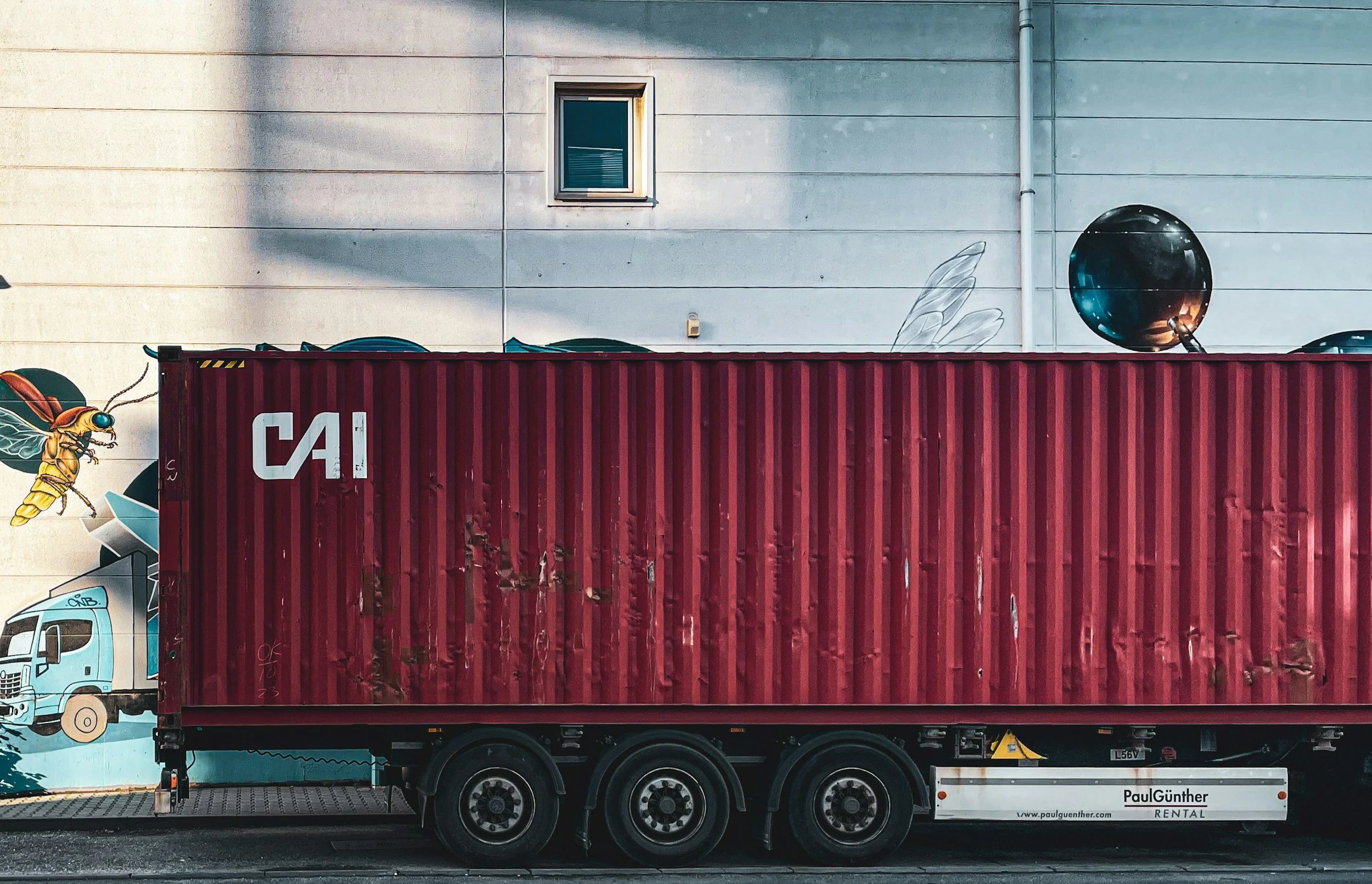 Red shipping container on a truck in front of a beige building with a small window and graffiti art on the wall.