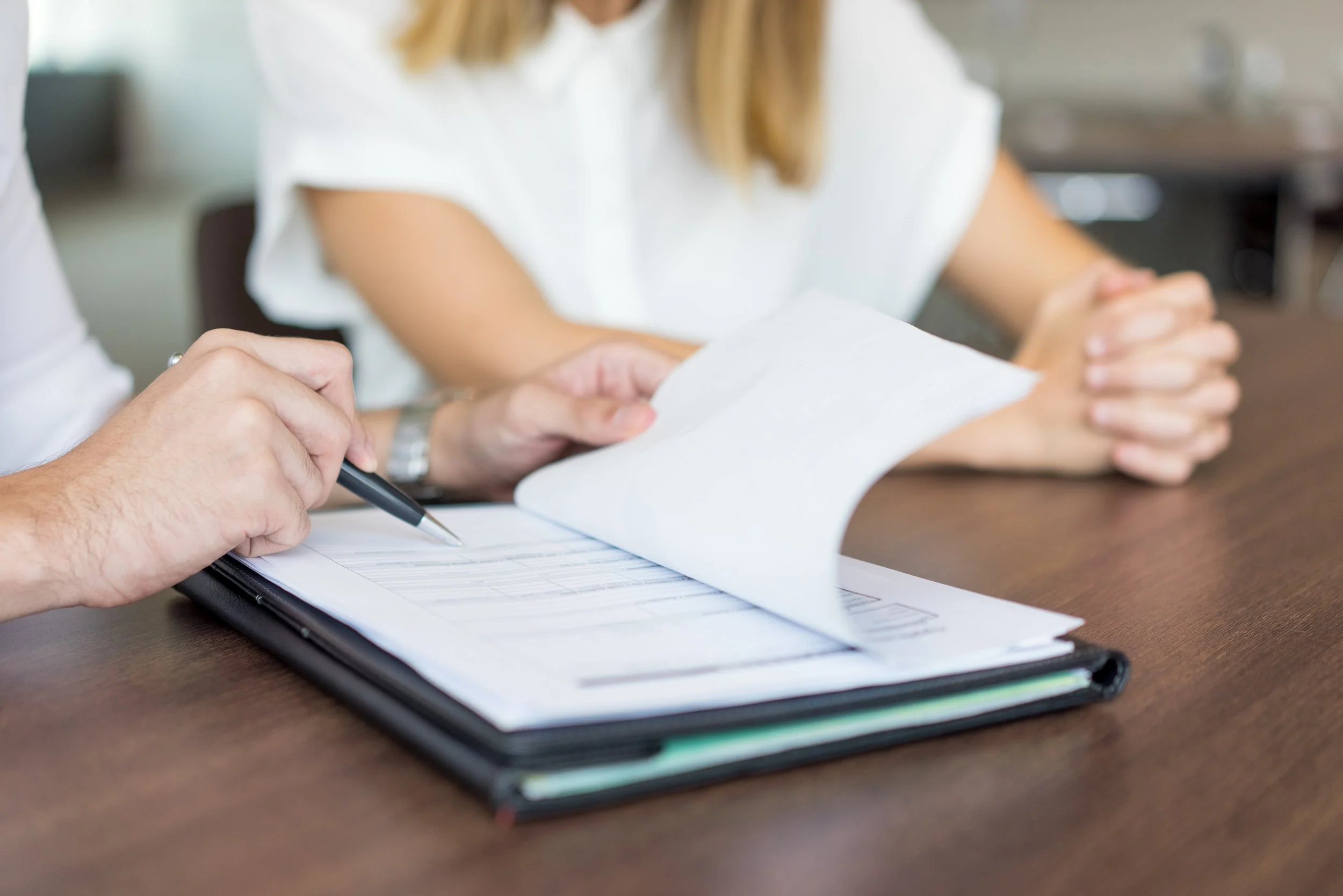 Close-up of a person's hand holding a pen and filling out a form on a clipboard, with another person's hands and folded hands resting on the table nearby.