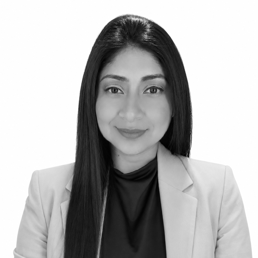Black and white photo of a woman with long dark hair, smiling, wearing a blazer, a black top, and a necklace with an apple-shaped pendant, standing against a plain background.