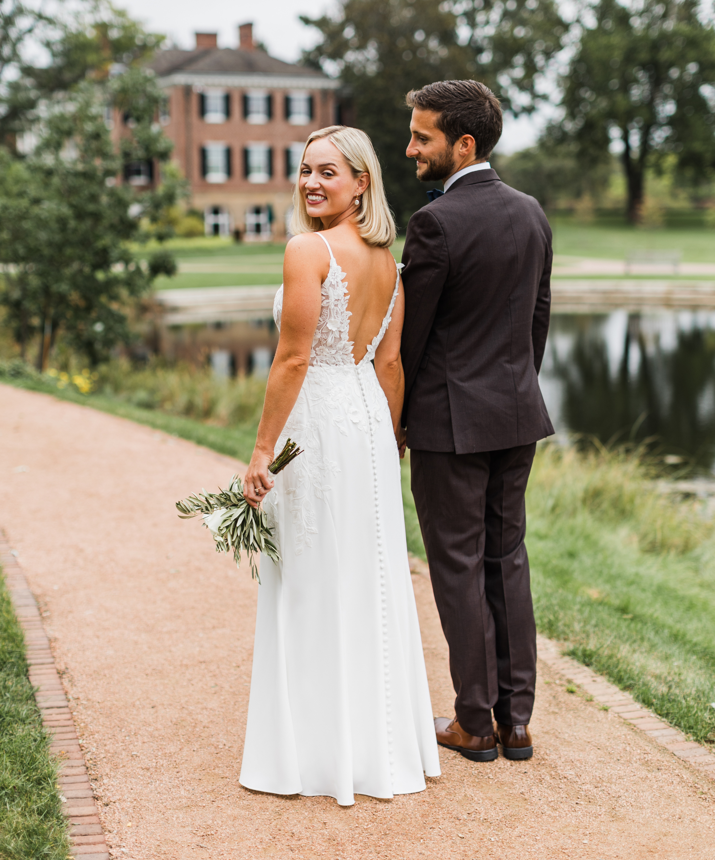Bride and groom standing on a path near a pond, holding hands, with a large historic brick house and trees in the background.