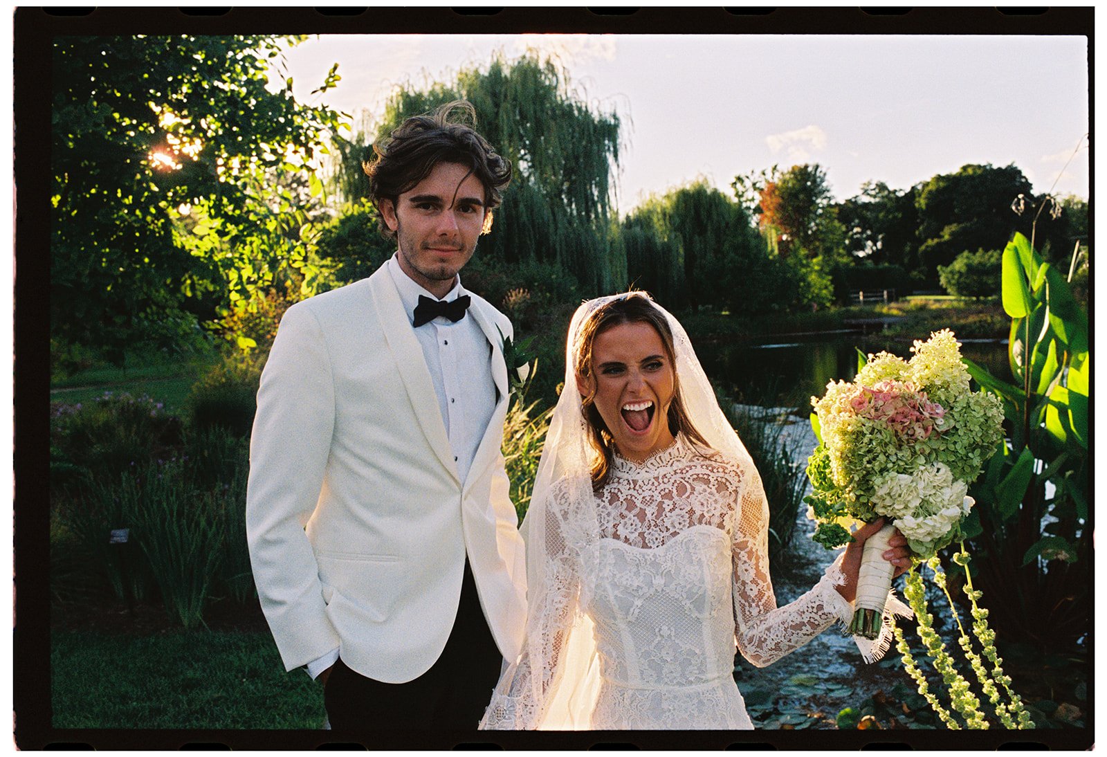 A bride in an all-lace wedding dress and a groom in a white jacket and black bowtie posing outside at the Cantigny East Lawn Garden, captured in film photography and managed by Chicago wedding planner One Fine Day Events.
