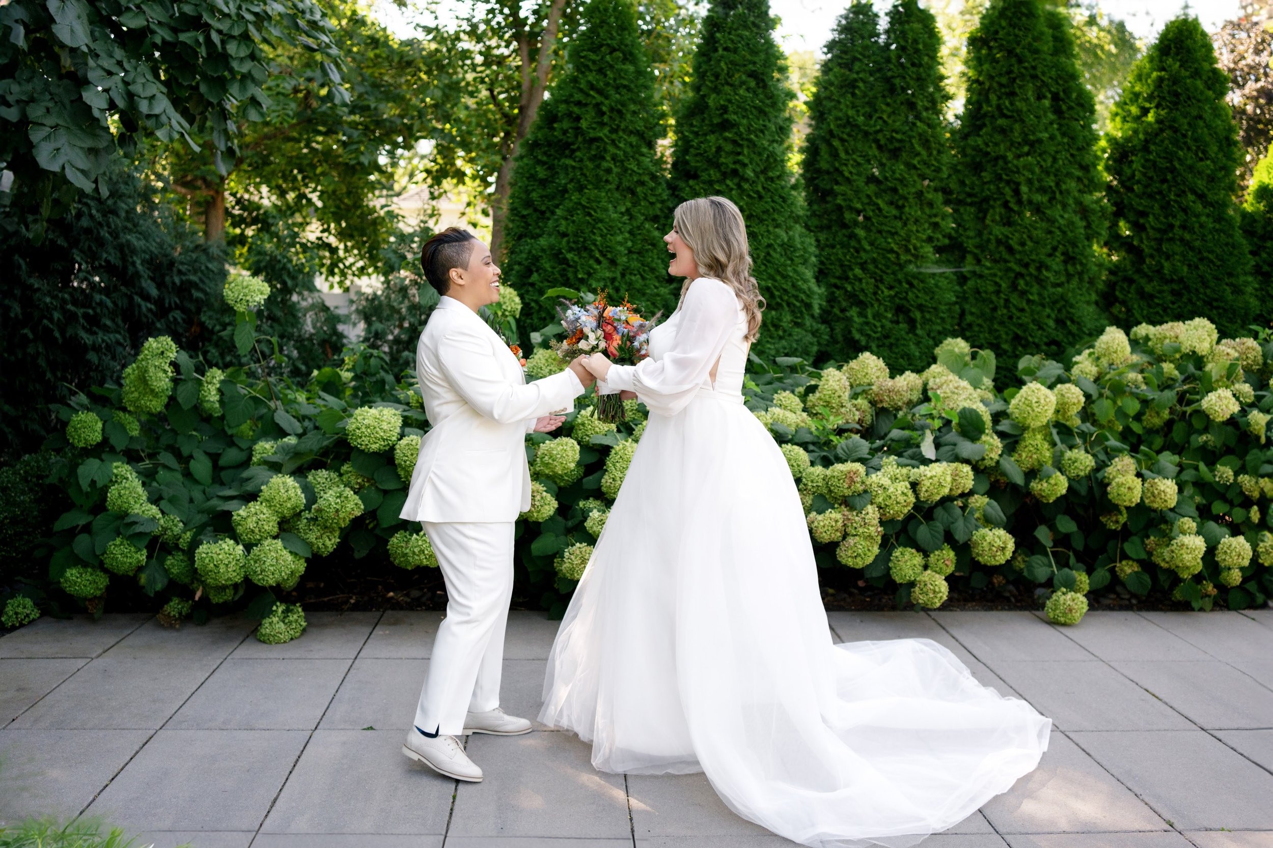 Two brides smiling outside their wedding ceremony at Warehouse 109 in Plainfield, IL; one bride in a white lace wedding dress and the other in a white tuxedo posing by a lush hydrangea bush, with planning by One Fine Day Events.