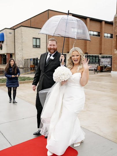 A bride and groom showing off their wedding rings while holding a clear umbrella at Sky on Nine in Rosemont, Illinois, during a rain-planned wedding day managed by One Fine Day Events.
