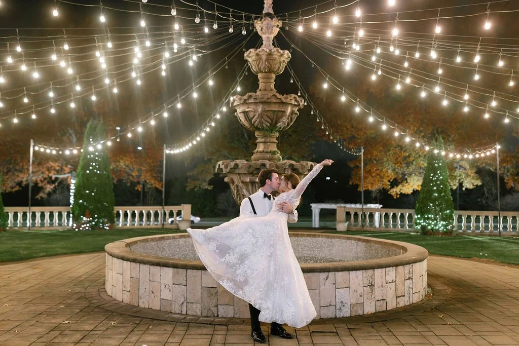 An excited bride and groom celebrating at night outside the fountain at The Drake Oak Brook; the groom in suspenders and a bow tie dips and kisses the bride as she raises her hand in joy, with planning by One Fine Day Events.