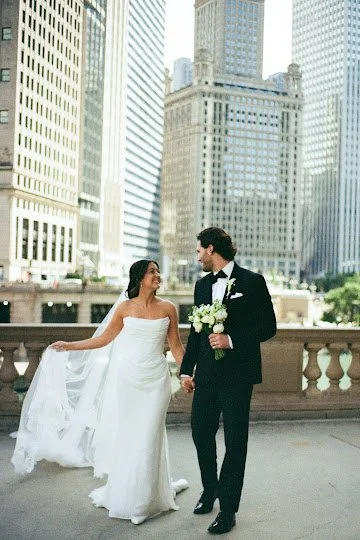 A bride in a strapless dress holding up her gown while the groom holds her white bouquet and hand, smiling at each other outside on the Chicago River during a stress-free city wedding planned by One Fine Day Events.