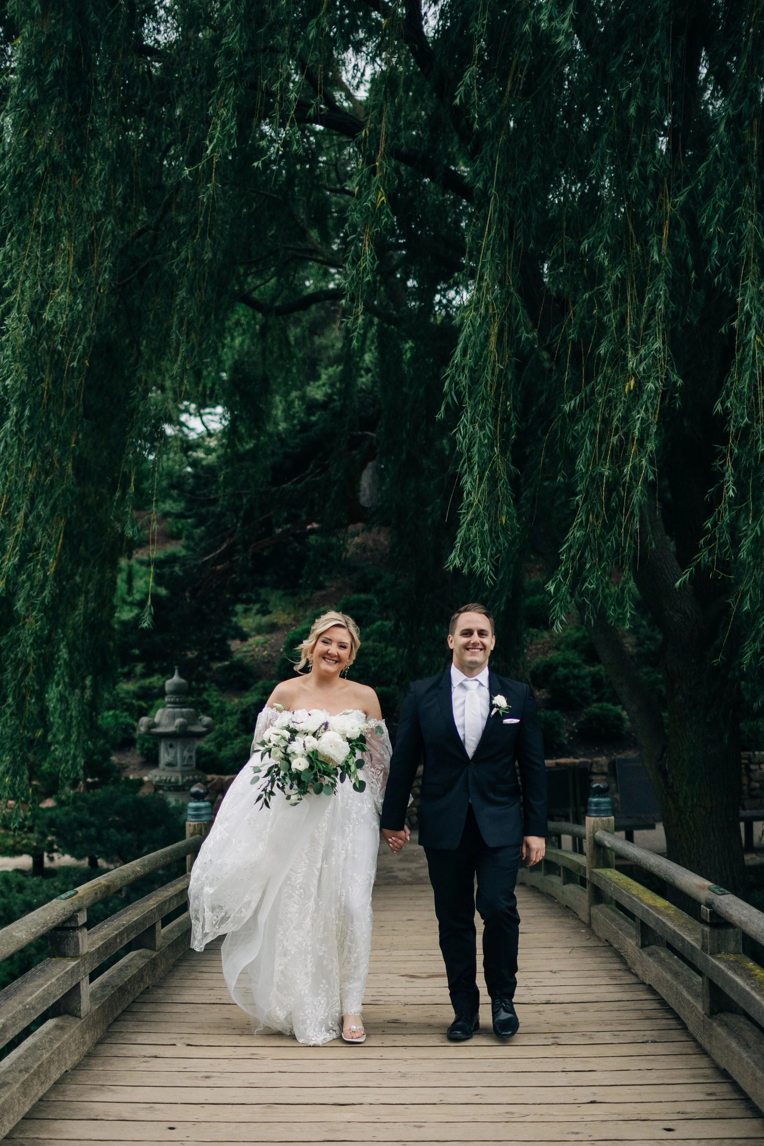Bride and groom walking hand in hand on a wooden bridge in a lush, green garden, smiling happily.