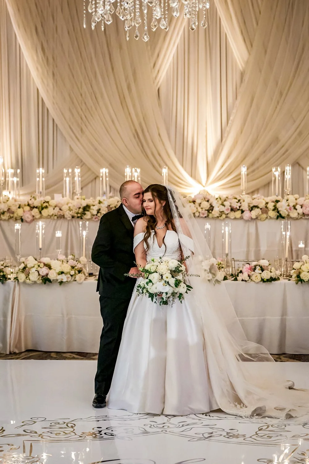 An exquisite bride being kissed by her groom at a luxury wedding head table featuring lush florals, elegant fabric draping, and a sea of glowing candles, designed and planned by Chicago wedding planner One Fine Day Events."