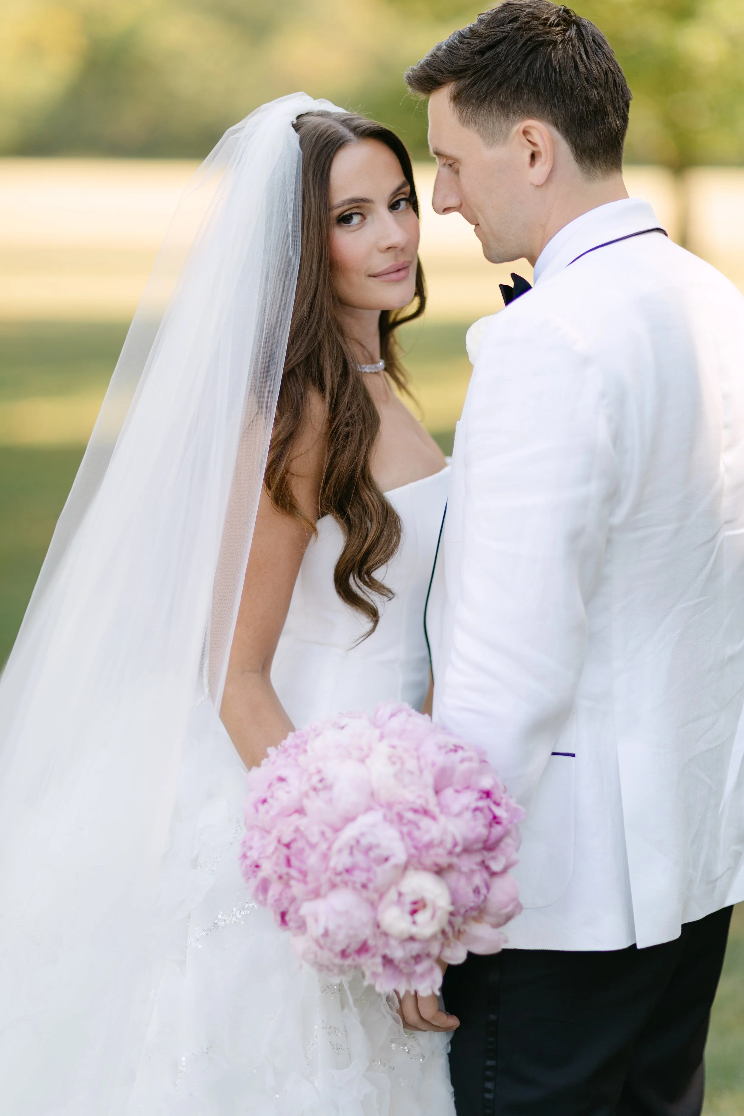 A beautiful bride in a strapless wedding gown with her hair down under a veil, holding a pink peony bouquet and posing with her groom in a white tuxedo jacket during a stress-free Chicago wedding planned by Chicago wedding planner One Fine Day Events