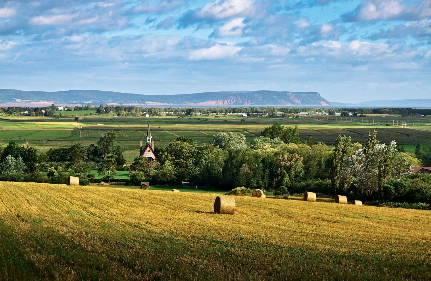 Rural landscape with hay bales, a church, and distant mountains under a cloudy sky.