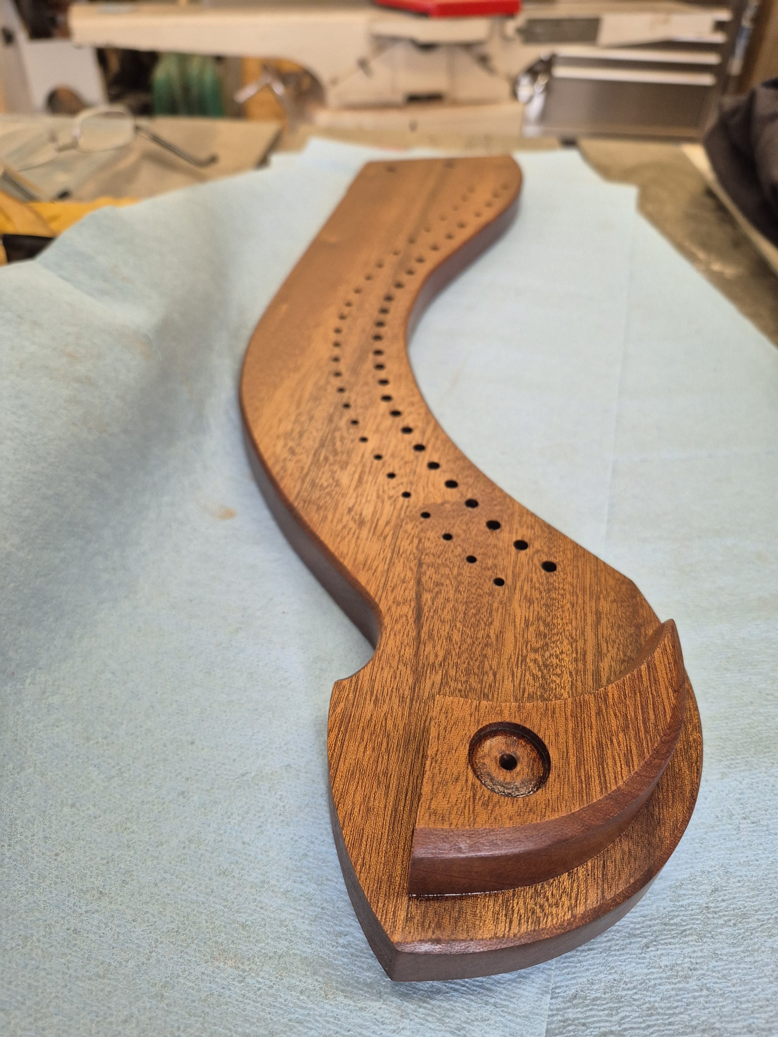 Close-up of a wooden cribbage board with a curved design, featuring a series of evenly spaced holes, placed on a soft surface in a workshop setting.