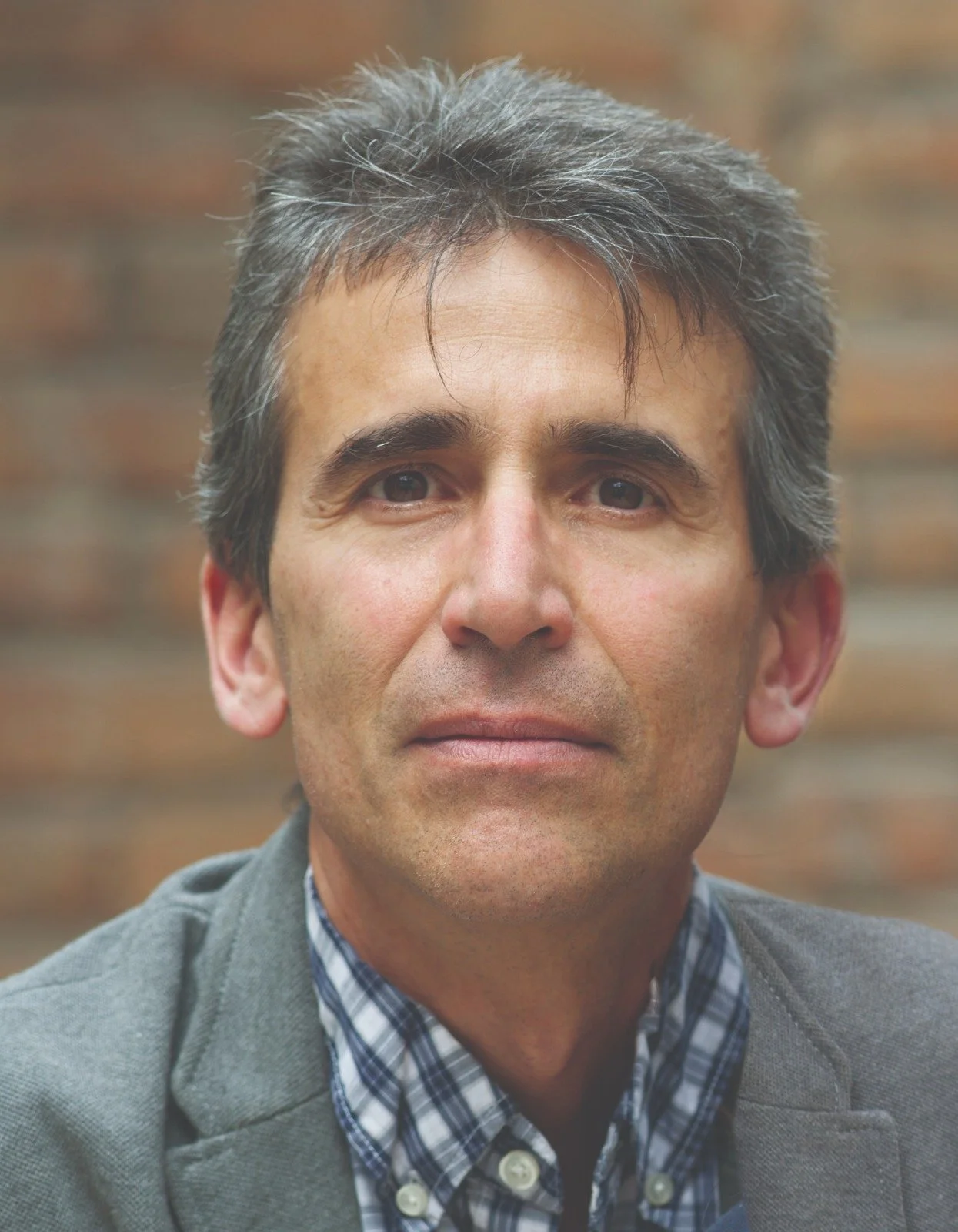 Retrato de Adolfo Macías Huerta con traje gris, camisa a cuadros en tonos azul y blanco, fondo de ladrillos.