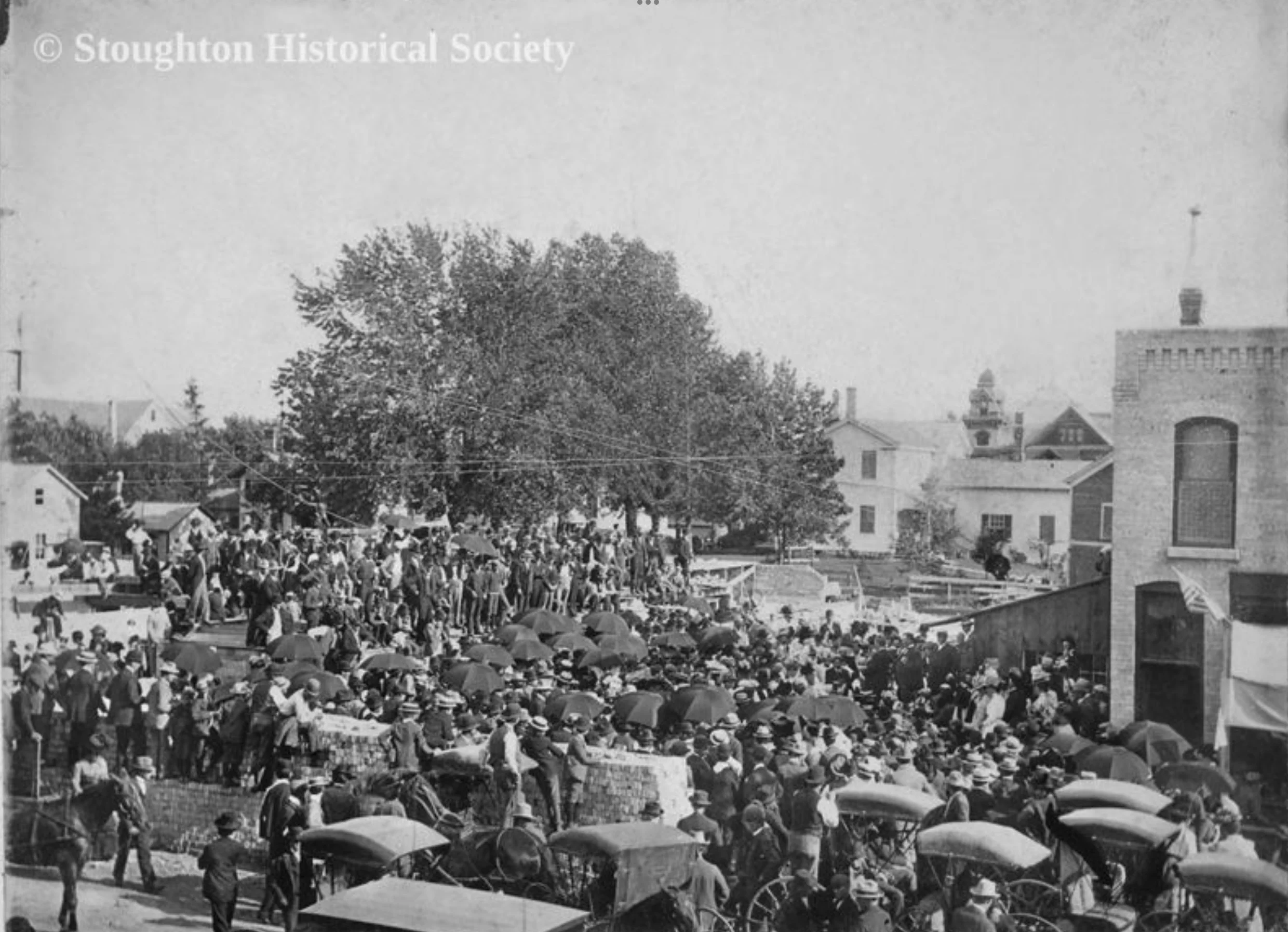 A black and white historical photo showing a large crowd gathered outdoors, many holding umbrellas, in front of the Stoughton Firehouse and City Hall.  There are buildings, trees, and power lines visible in the background.