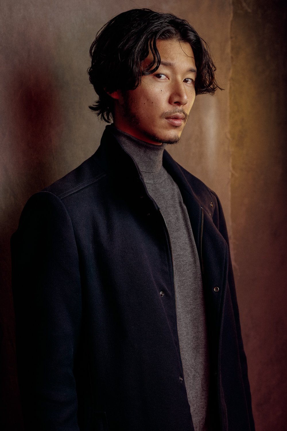 Portrait of a man with medium-length dark hair, wearing a dark coat and gray turtleneck, in a warm-toned setting.
