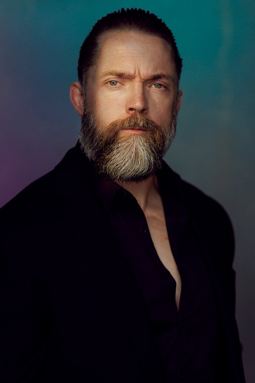 Man with beard and stern expression wearing a dark suit against a colorful background.