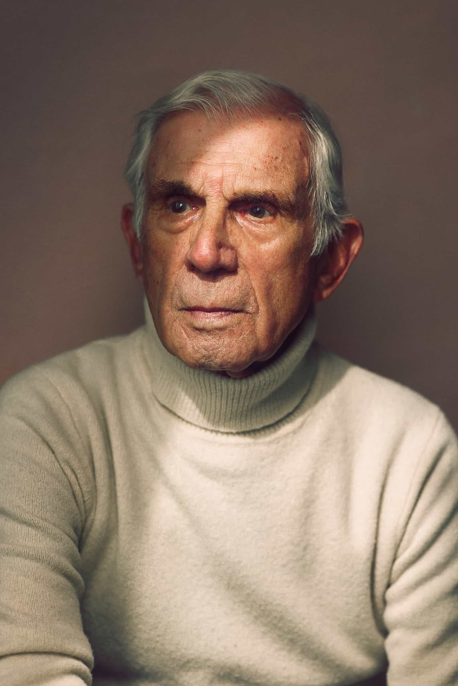 Elderly man wearing a beige turtleneck sweater, looking thoughtful.