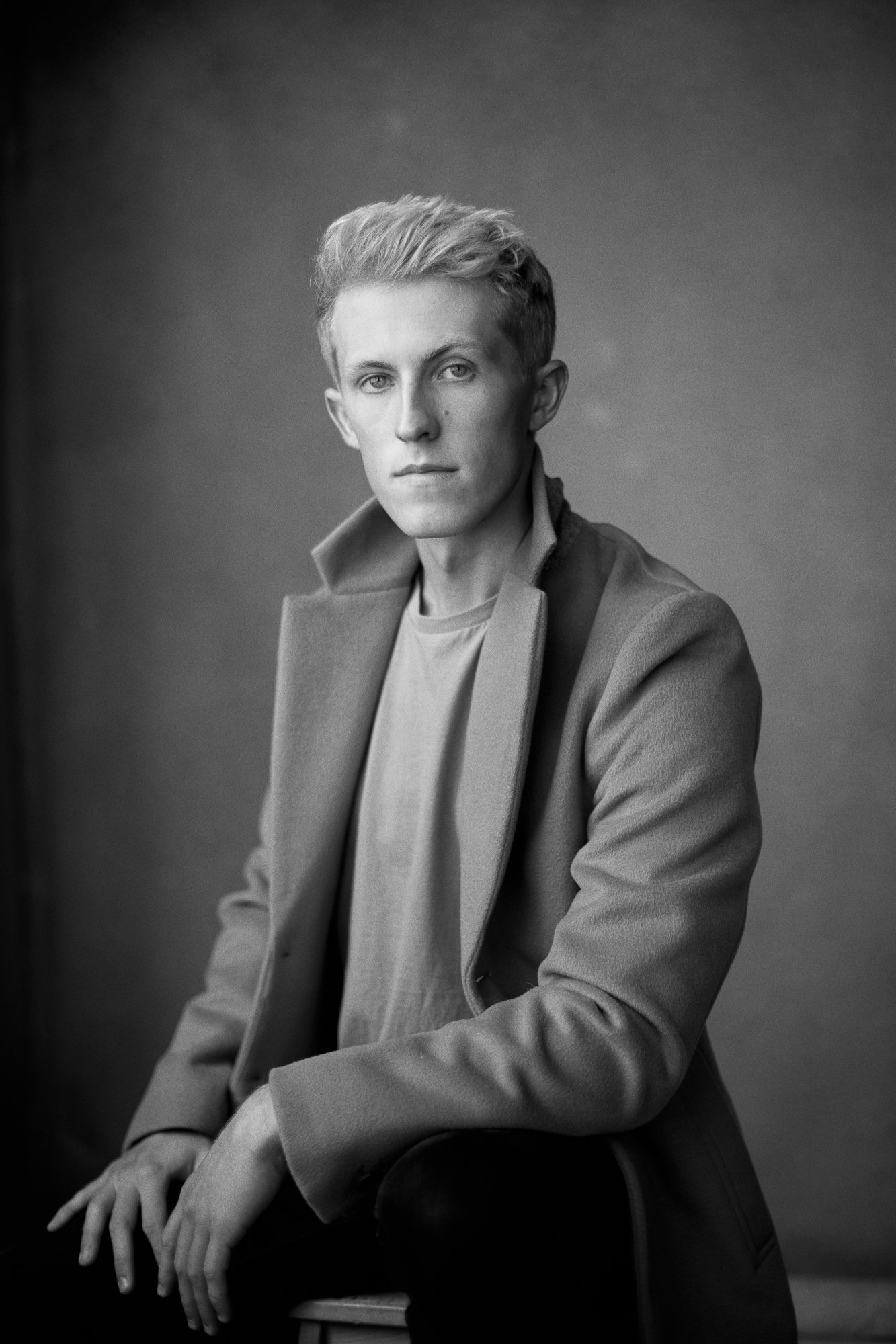 Black and white portrait of a person in a coat sitting on a stool, looking at the camera.