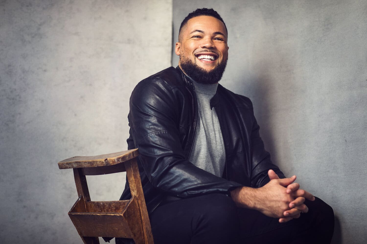A man sitting and smiling, wearing a leather jacket and gray shirt, seated on a wooden chair.