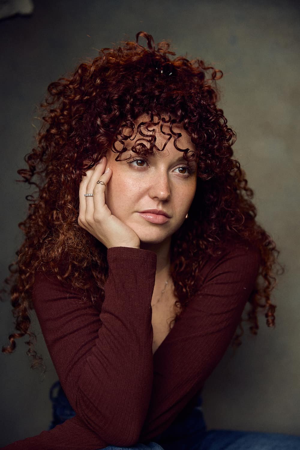 Woman with curly hair wearing a brown long-sleeve shirt, resting her chin on her hand.
