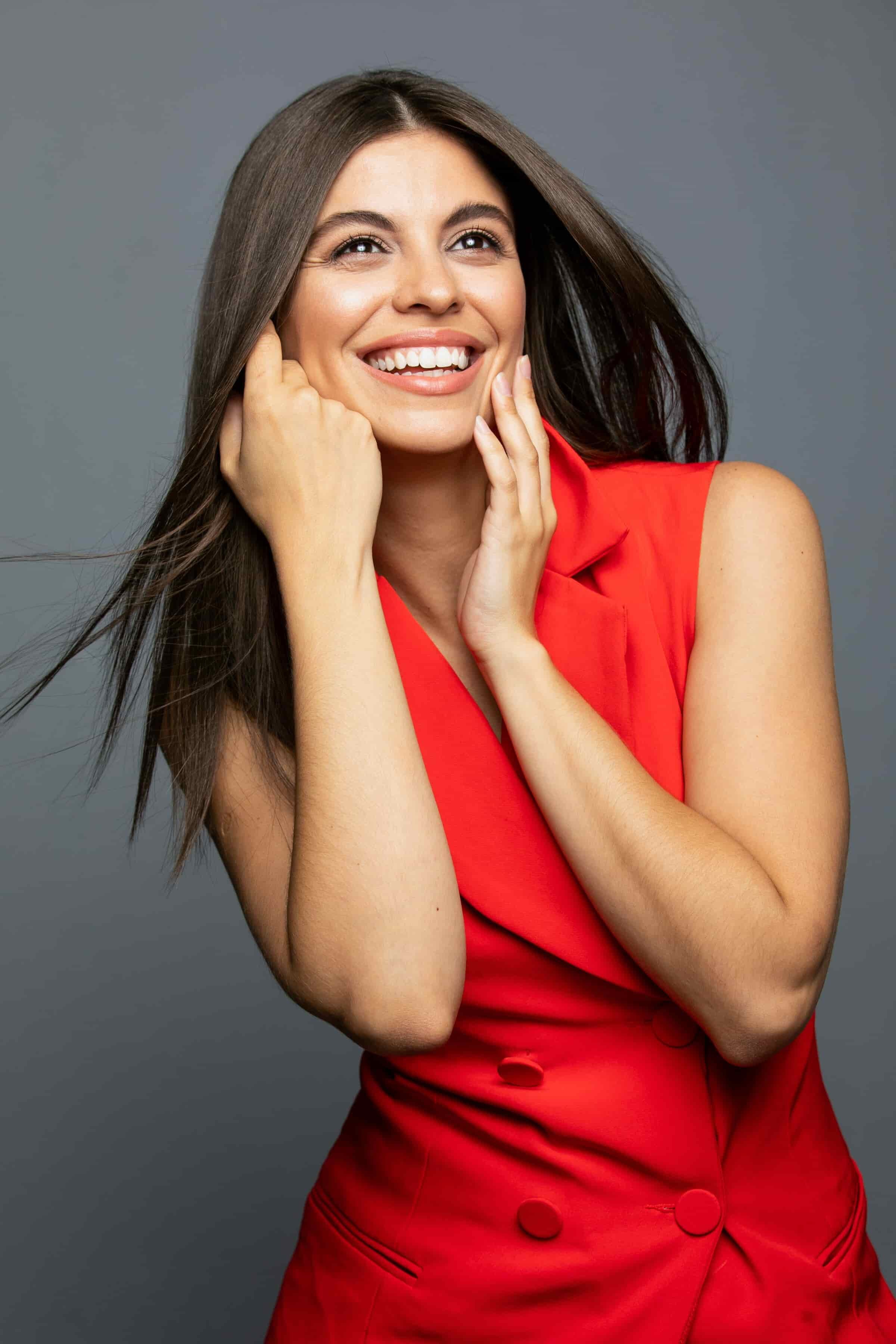 A woman smiling, wearing a red sleeveless top, with long brown hair against a gray background.