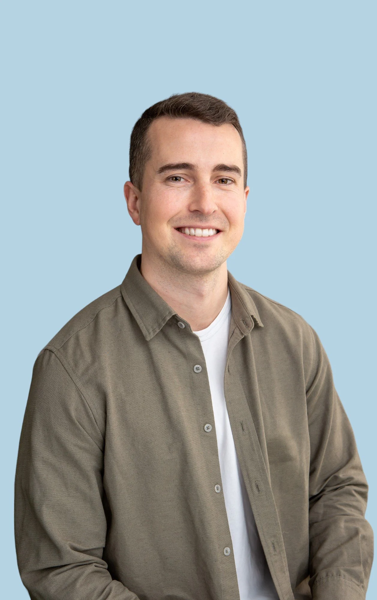 Smiling person wearing a beige shirt and white t-shirt against a light blue background.