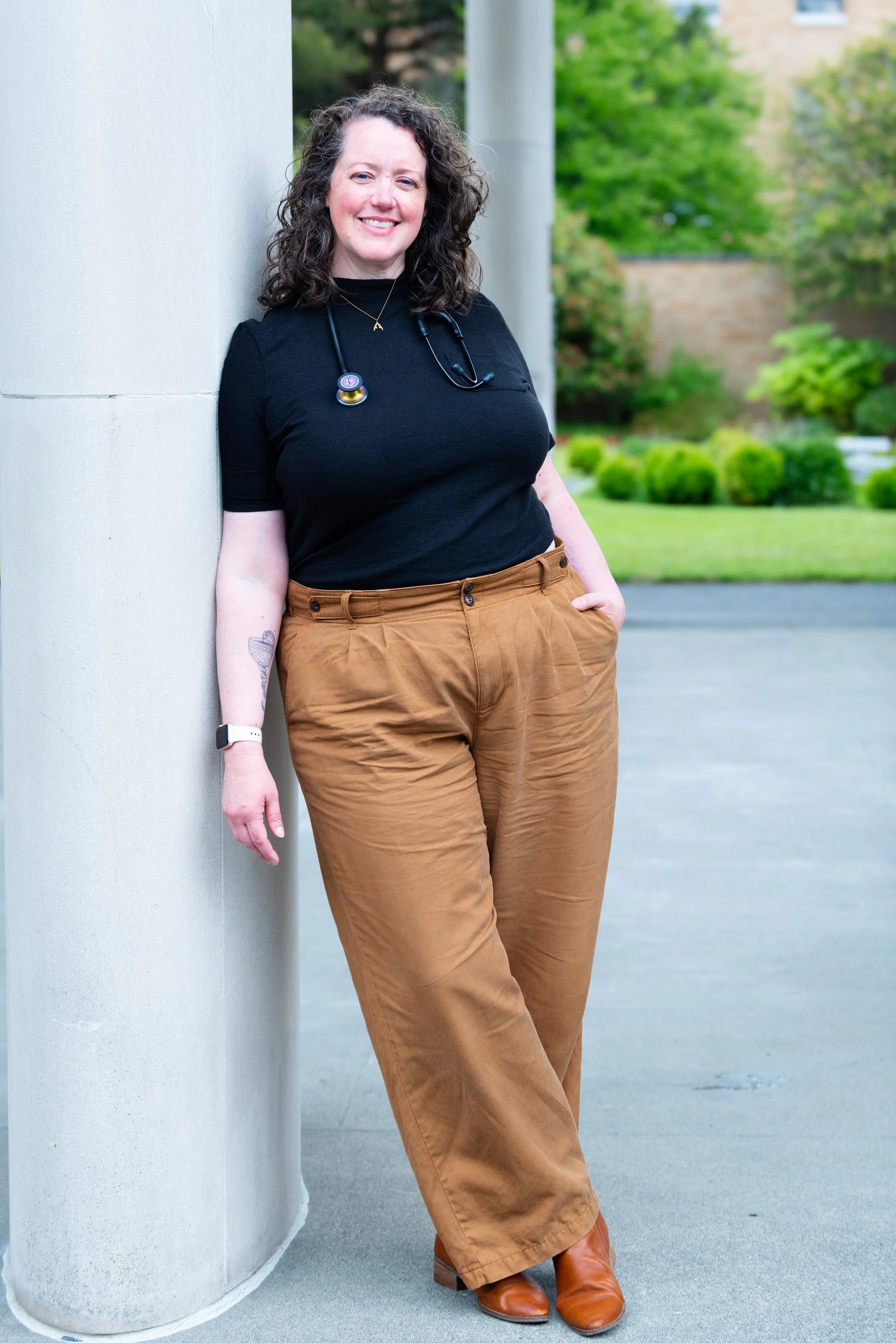 A smiling woman with curly hair, wearing a black top, brown pants, brown boots, and a stethoscope around her neck, leaning against a large grey pillar outdoors with green trees and bushes in the background.