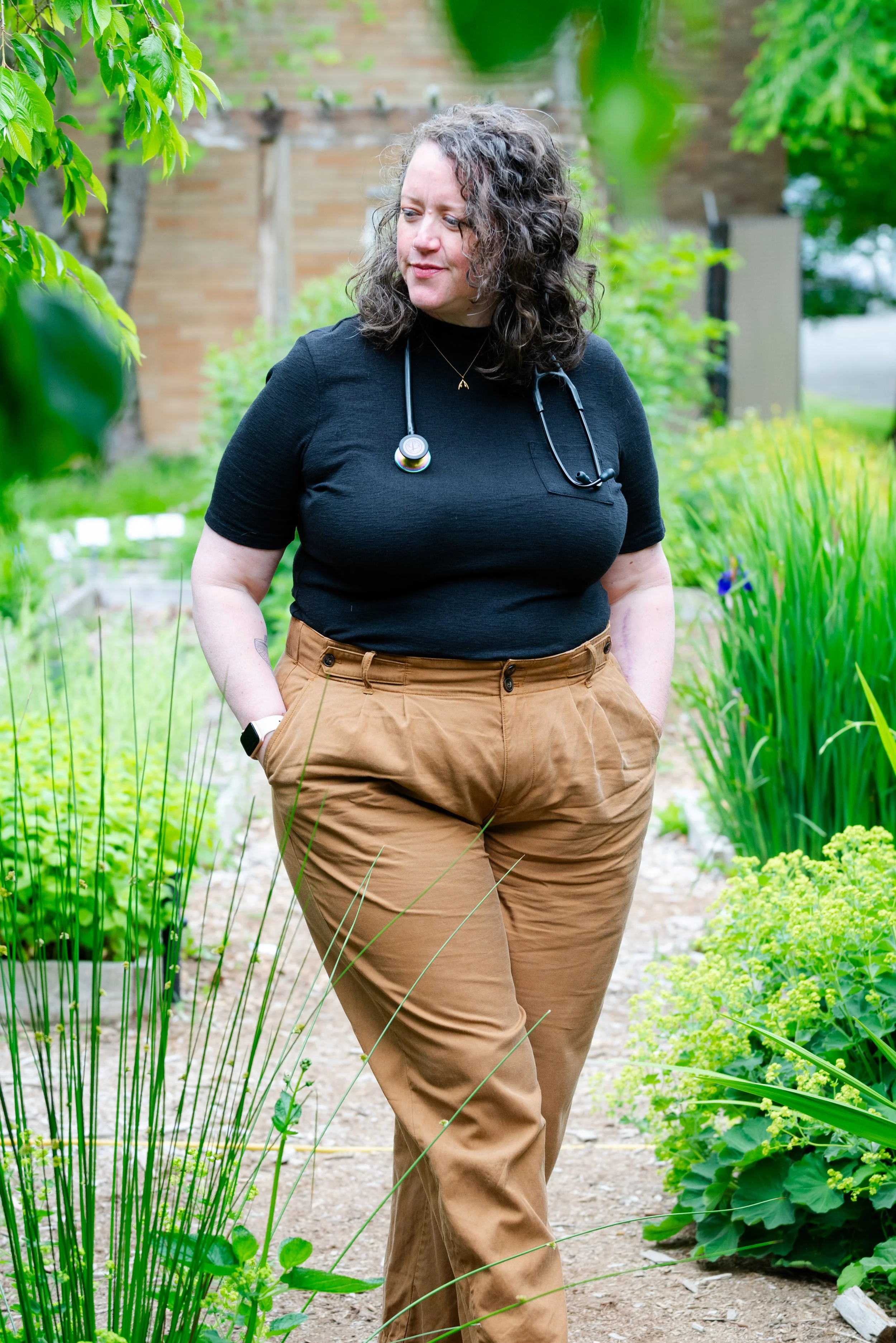 A woman with curly hair and a stethoscope around her neck walking in a lush garden with green plants.
