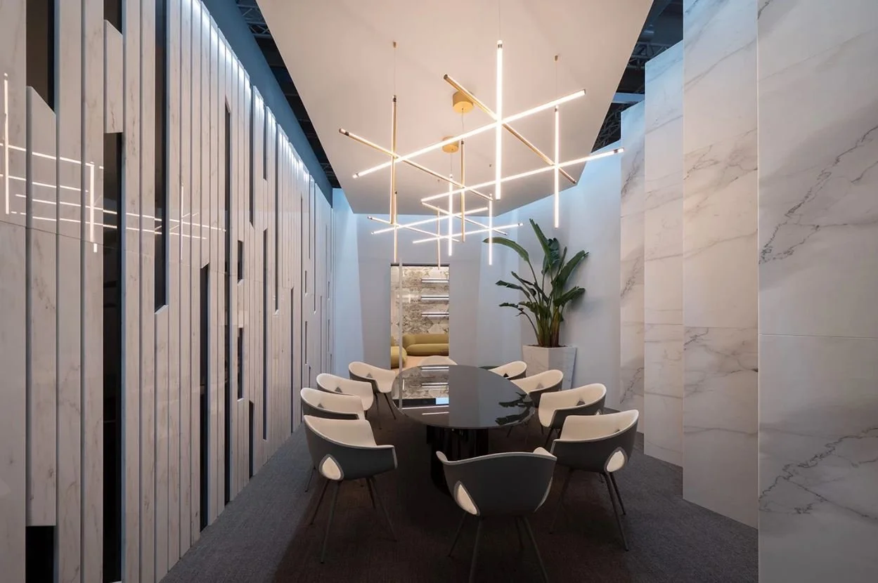 Interno di una sala conferenze moderna con tavolo ovale nero circondato da sedie bianche e grigie, illuminazione a sospensione stilizzata e pareti decorate con pannelli effetto marmo. Un grande vaso con pianta verde è visibile sullo sfondo. arch-12 A