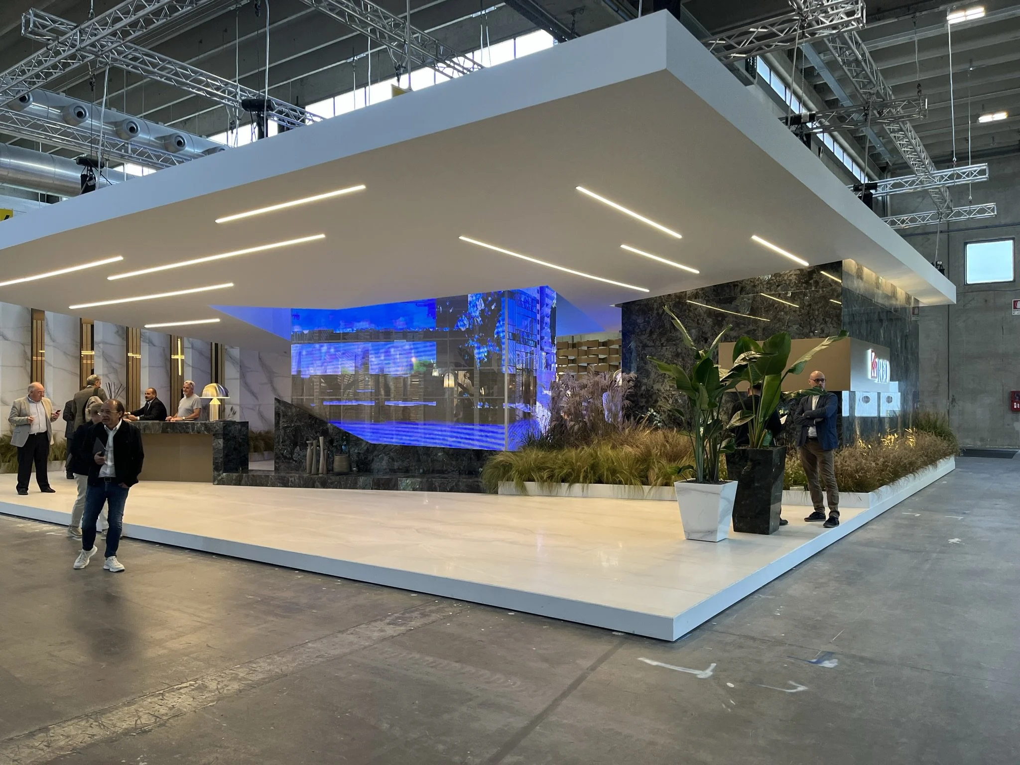 Esposizione in fiera con stand moderno e design elegante, persone che interagiscono, piante decorative e luci integrate nel soffitto. arch-12 A-12  Simone Rosi design