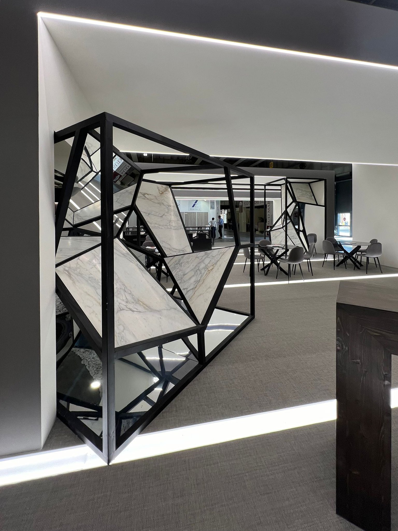 Interno di uno spazio espositivo moderno con struttura geometrica in metallo e marmo, con tavoli e sedie nell'area sullo sfondo. arch-12 A-12 Simone Rosi design