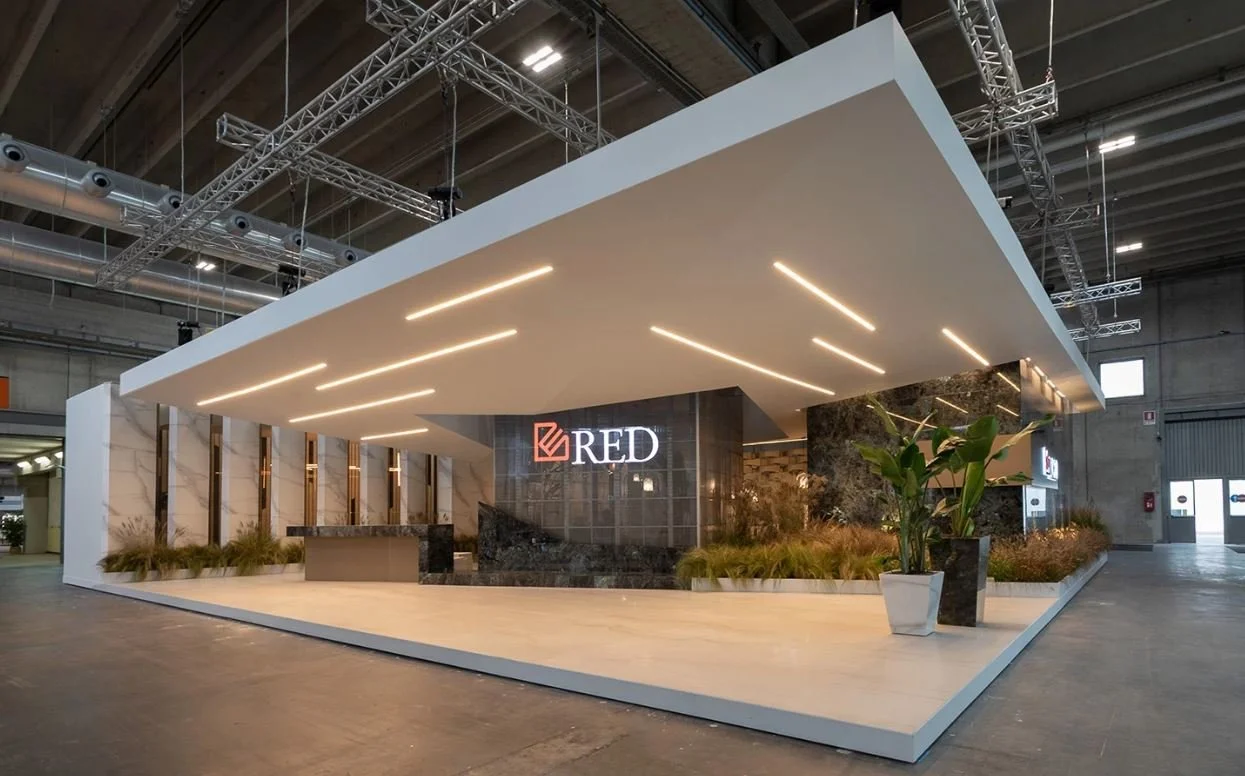 Un moderno stand espositivo in un edificio industriale con illuminazione elegante e piante decorative. Un grande logo "RED" è visibile sulla struttura. La pavimentazione è chiara e liscia, e l'ambiente è spazioso con un soffitto alto e strutture in m
