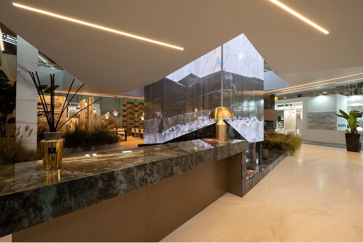 Interno di un'area espositiva moderna con bancone in marmo, lampada dorata, decori vegetali e luci a soffitto. Piante ornamentali e dettagli di design contemporaneo.arch-12 A-12  Simone Rosi design