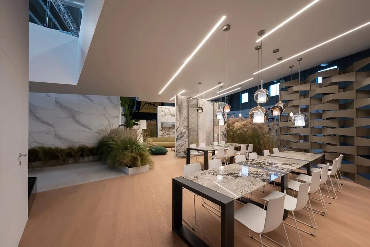 Interno di uno spazio espositivo moderno con tavoli in marmo, sedie bianche, lampade a sospensione e piante decorative. arch-12 A-12  Simone Rosi design