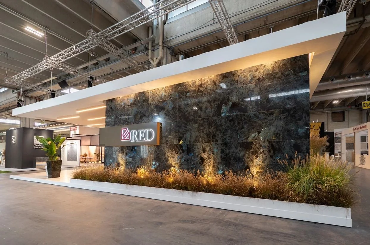 Stand fieristico aziendale con logo "RED" su parete con rivestimento scuro, decorato con piante e illuminazione soffusa, in un ambiente espositivo industriale. arch-12 A-12  Simone Rosi design