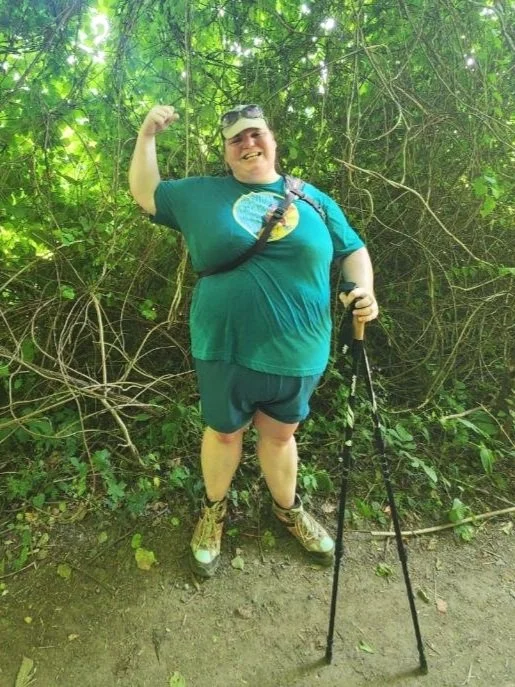 Woman smiling and flexing her arm in strength, standing in front of dense foliage, wearing a teal shirt, shorts, and hiking boots, holding trekking poles, with greenery surrounding them.