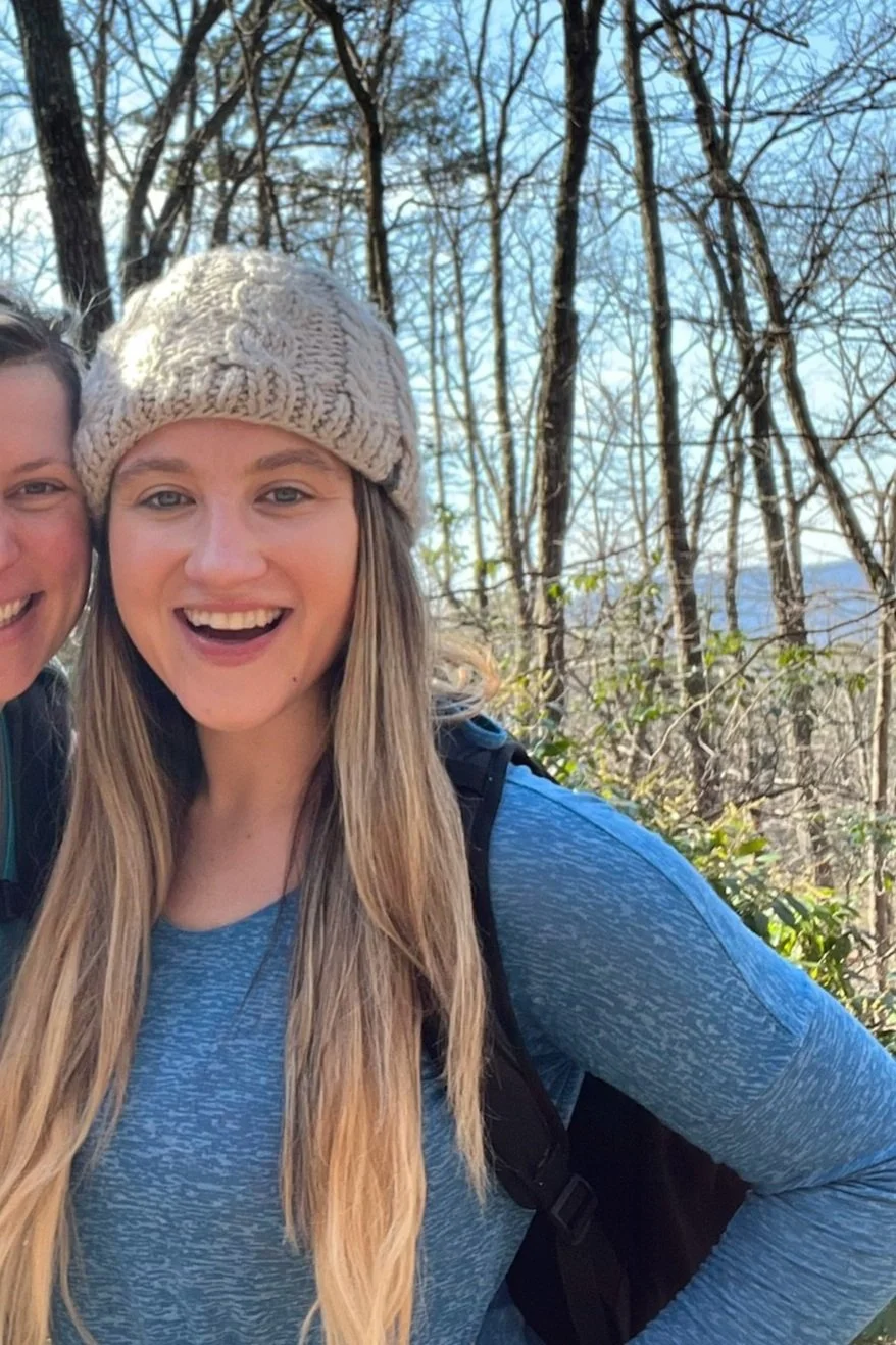 Woman wearing a beanie and blue long-sleeve shirt, smiling outdoors with trees in the background.