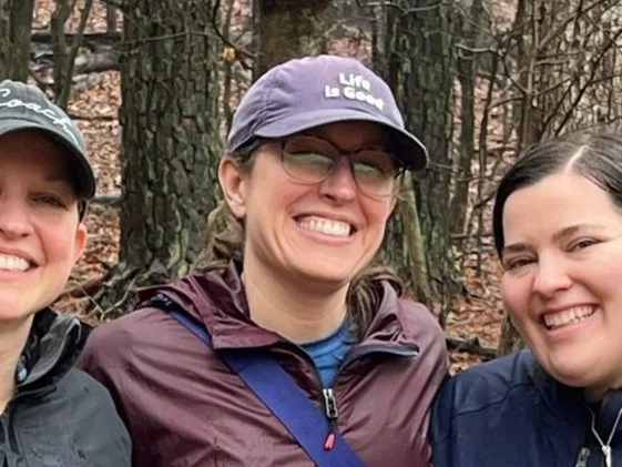 A woman in a baseball cap smiling in the woods in the fall