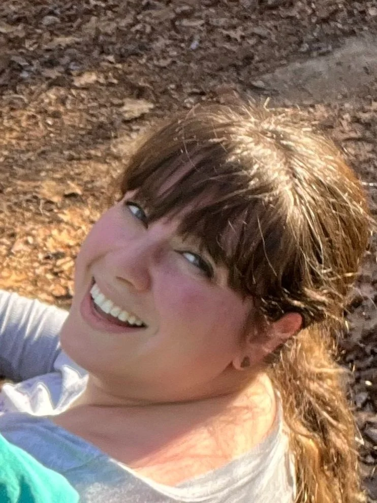 A woman smiling, lying on the ground covered with autumn leaves, wearing a gray shirt with sunlight on her hair.