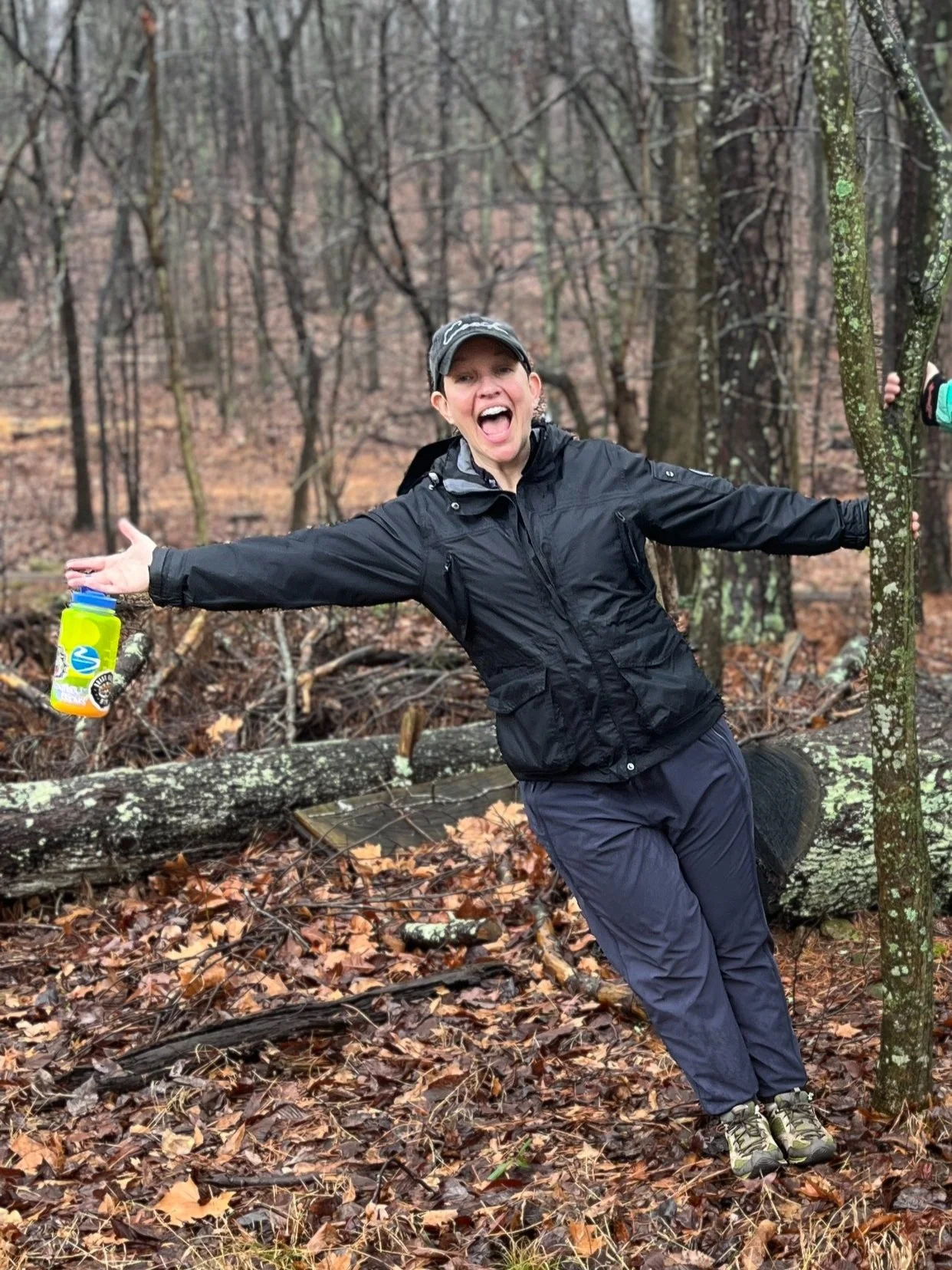 Person in a black jacket and hat smiling in a forest, holding a water bottle, surrounded by trees with fallen brown leaves on the ground.
