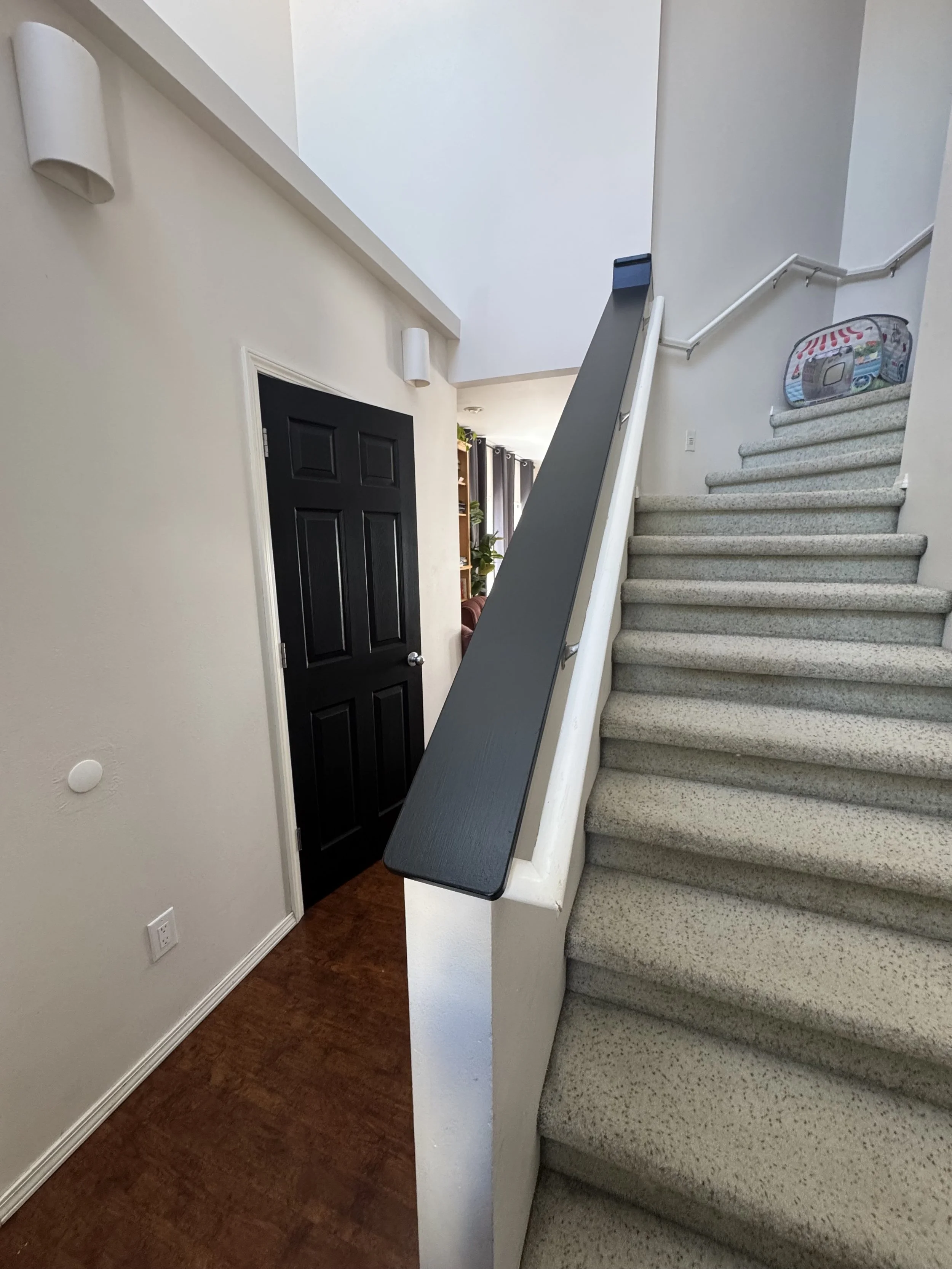 Interior view of a staircase with beige carpeted stairs, a black door on the left, a small potted plant, and a corner with a laundry basket with a colorful design at the top of the stairs.