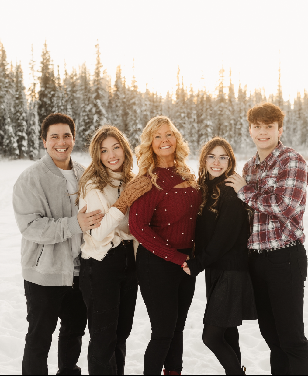 A family of five standing outdoors in snow with fir trees in the background, smiling and posing for a photo during winter.