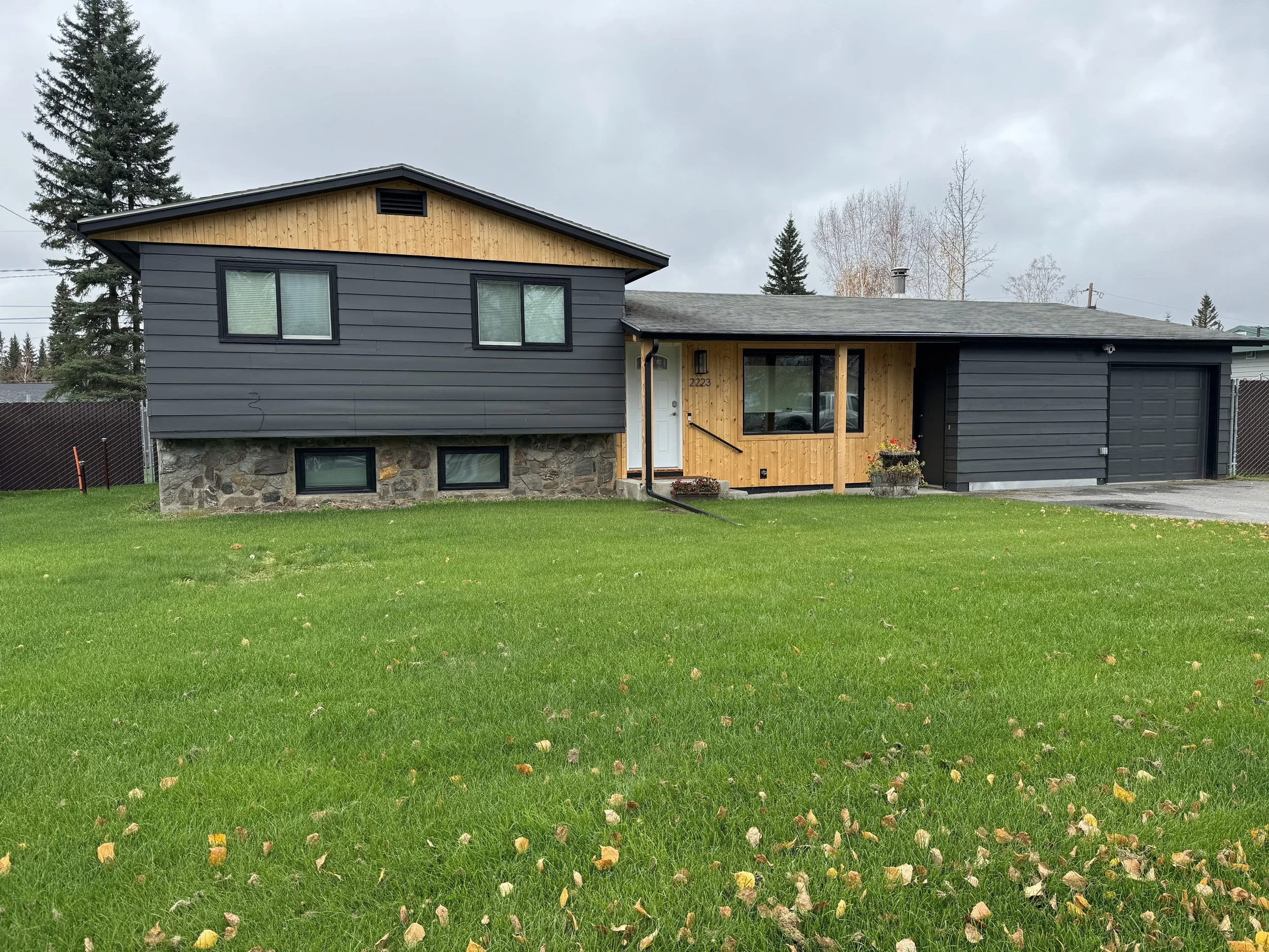 Front view of a house with dark gray siding and a wooden accent on the upper facade, a stone foundation, and a well-maintained lawn with fallen leaves. The house has two large windows in the upper section and two smaller basement windows. The sky is overcast with gray clouds, and there are tall trees visible in the background.