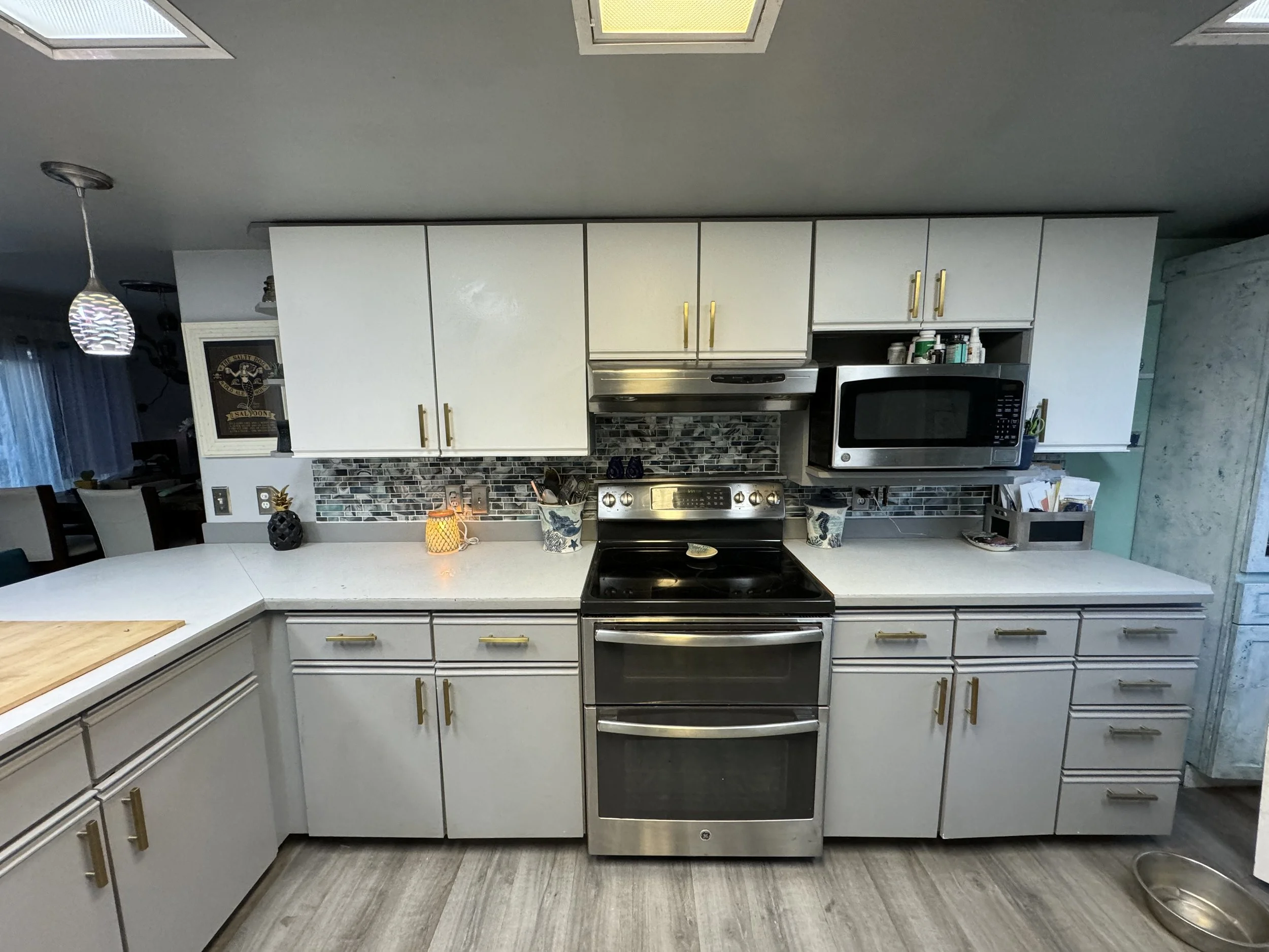 A modern kitchen with white cabinets, gold handles, a stainless steel oven, microwave, and a tiled backsplash. There are decorative items, including a candle, a potted plant, and kitchen utensils on the countertop.