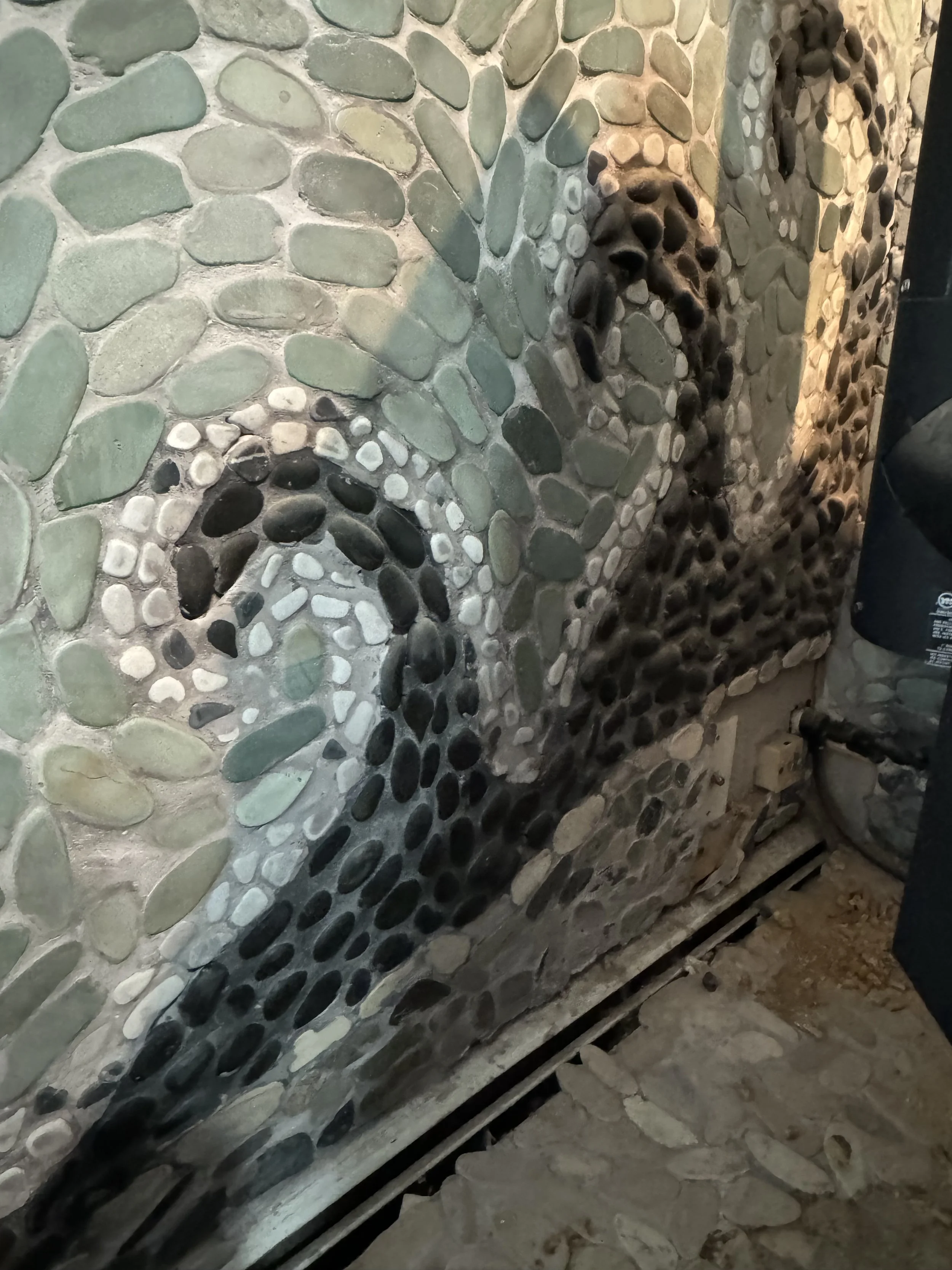 Close-up view of a stone wall with rounded, multicolored pebbles arranged in decorative patterns, with a section of the supporting dirt ground at the bottom.