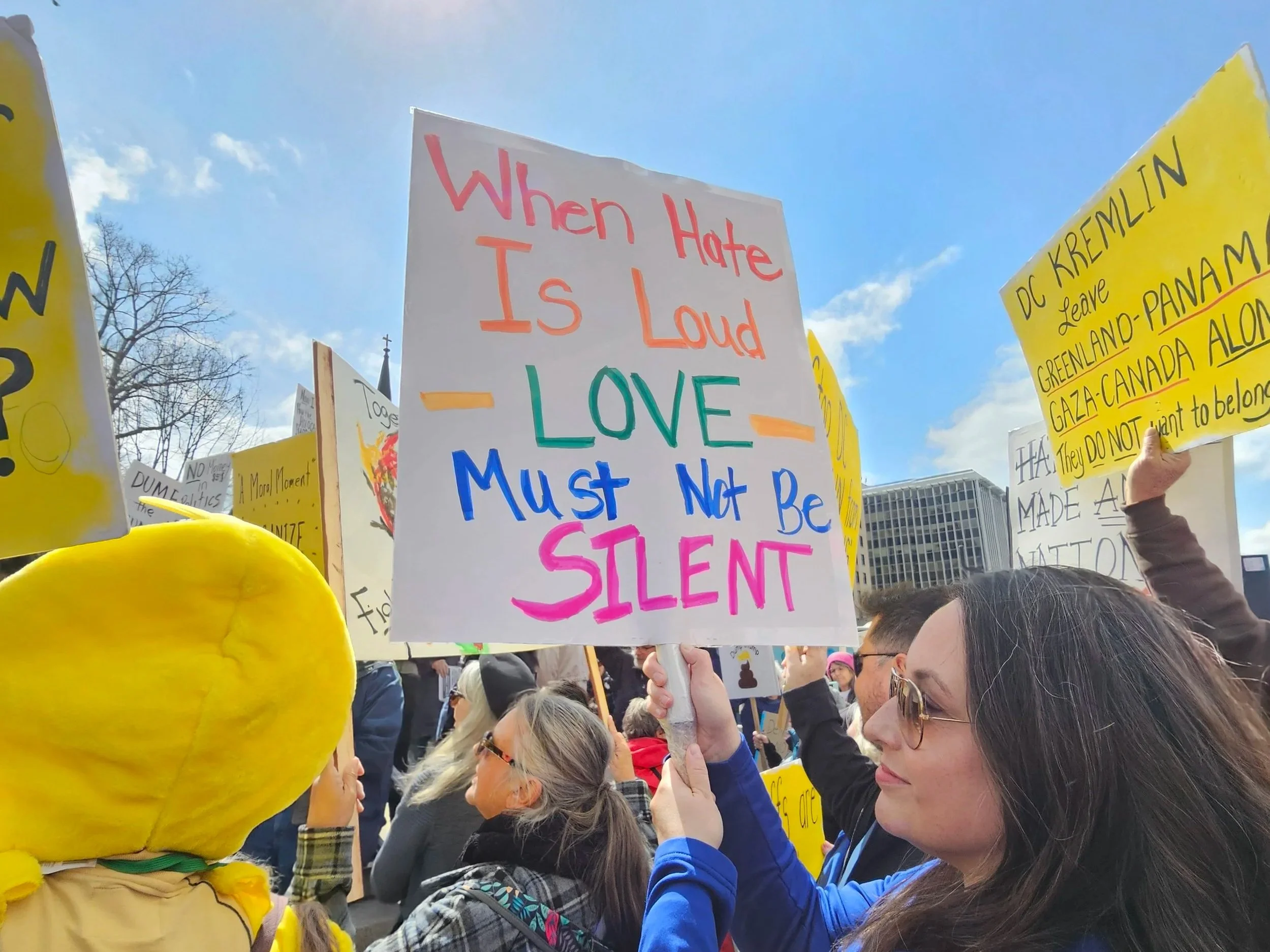 A woman holding a sign that reads, "When hate is loud, love must not be silent," at a protest or rally outdoors with other people holding signs under a sunny sky.