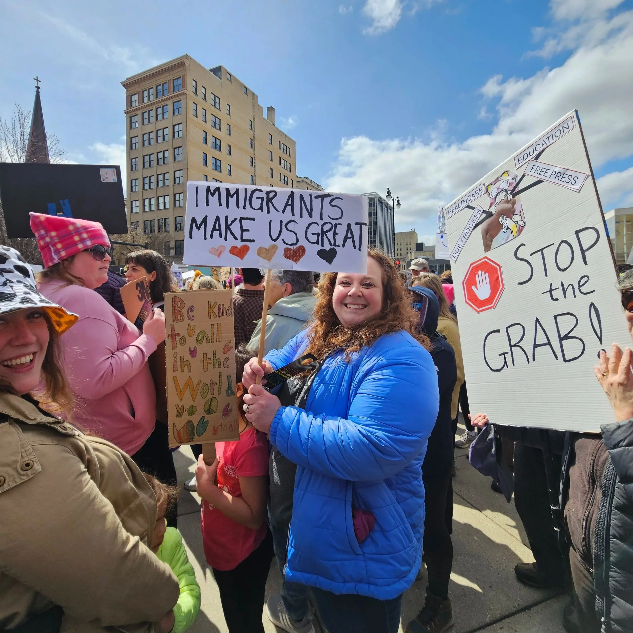 A woman in a blue jacket smiling at a protest holding a sign that says, 'Immigrants make us great'. Other protesters with signs include one with 'Stop the grab' and another with a message about kindness and diversity. The crowd is gathered outdoors i