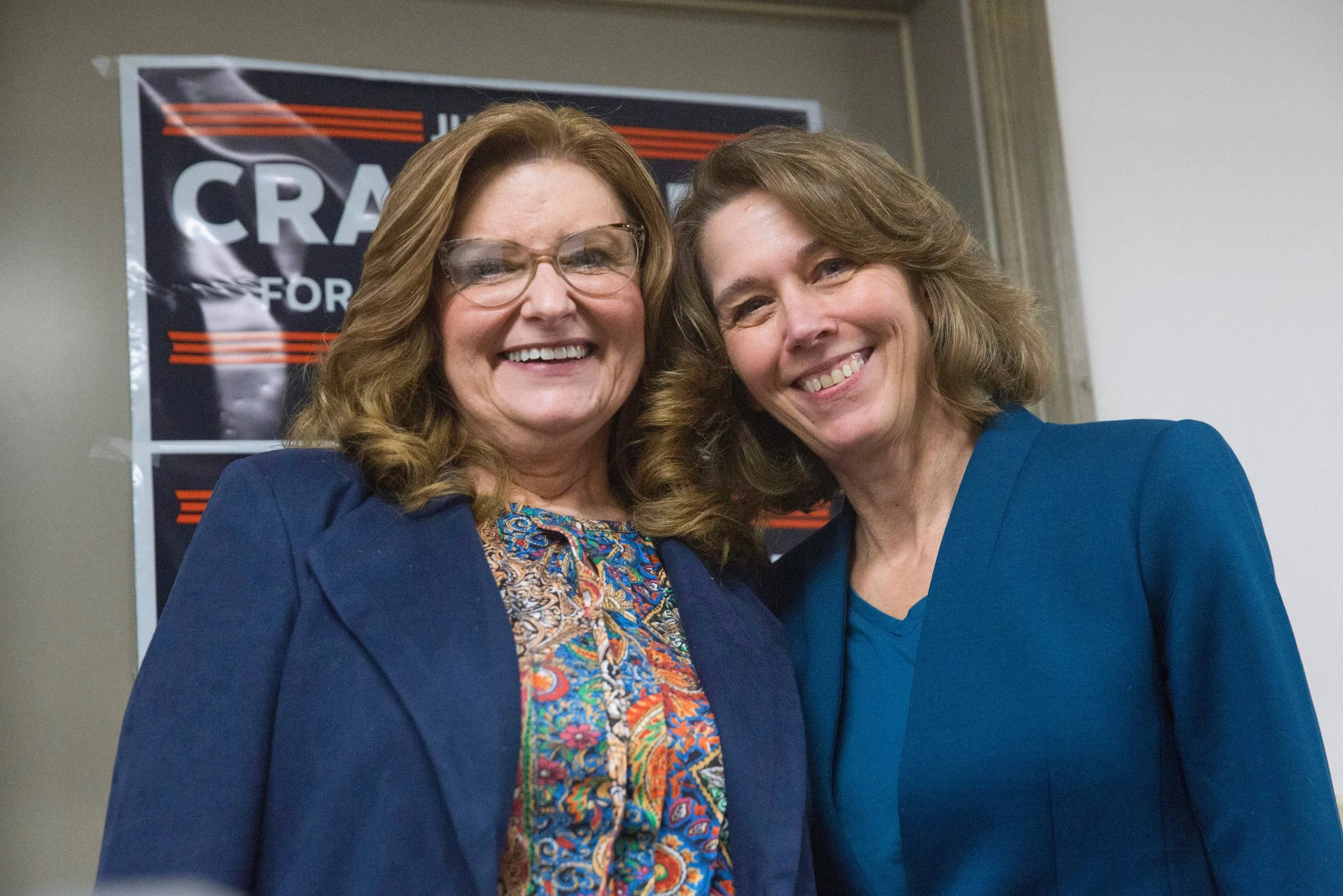 Two smiling women standing together in front of a campaign poster, dressed in blue blazers, in an indoor setting.