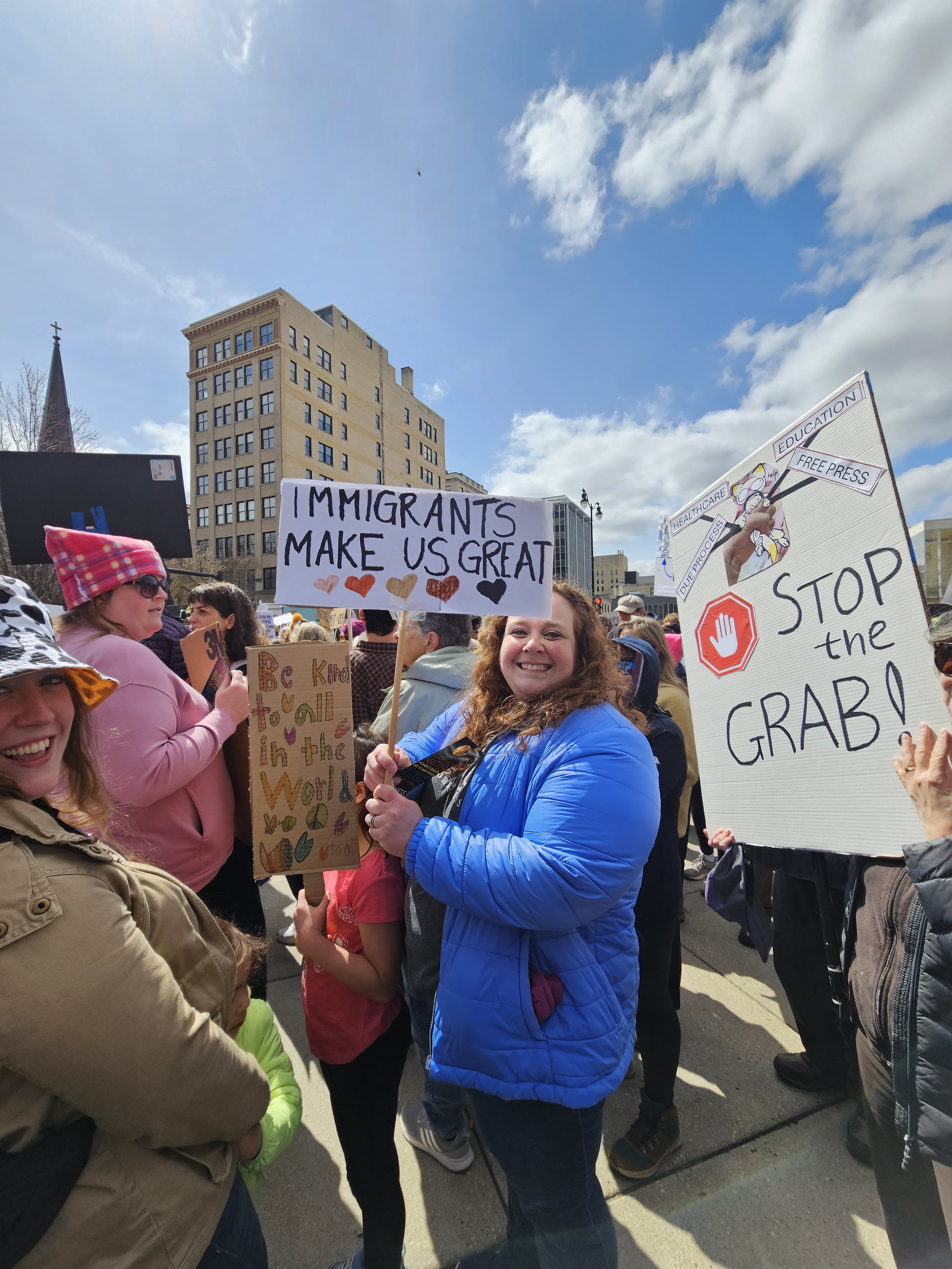 A woman in a blue jacket smiling at a protest holding a sign that says, 'Immigrants make us great'. Other protesters with signs include one with 'Stop the grab' and another with a message about kindness and diversity. The crowd is gathered outdoors i