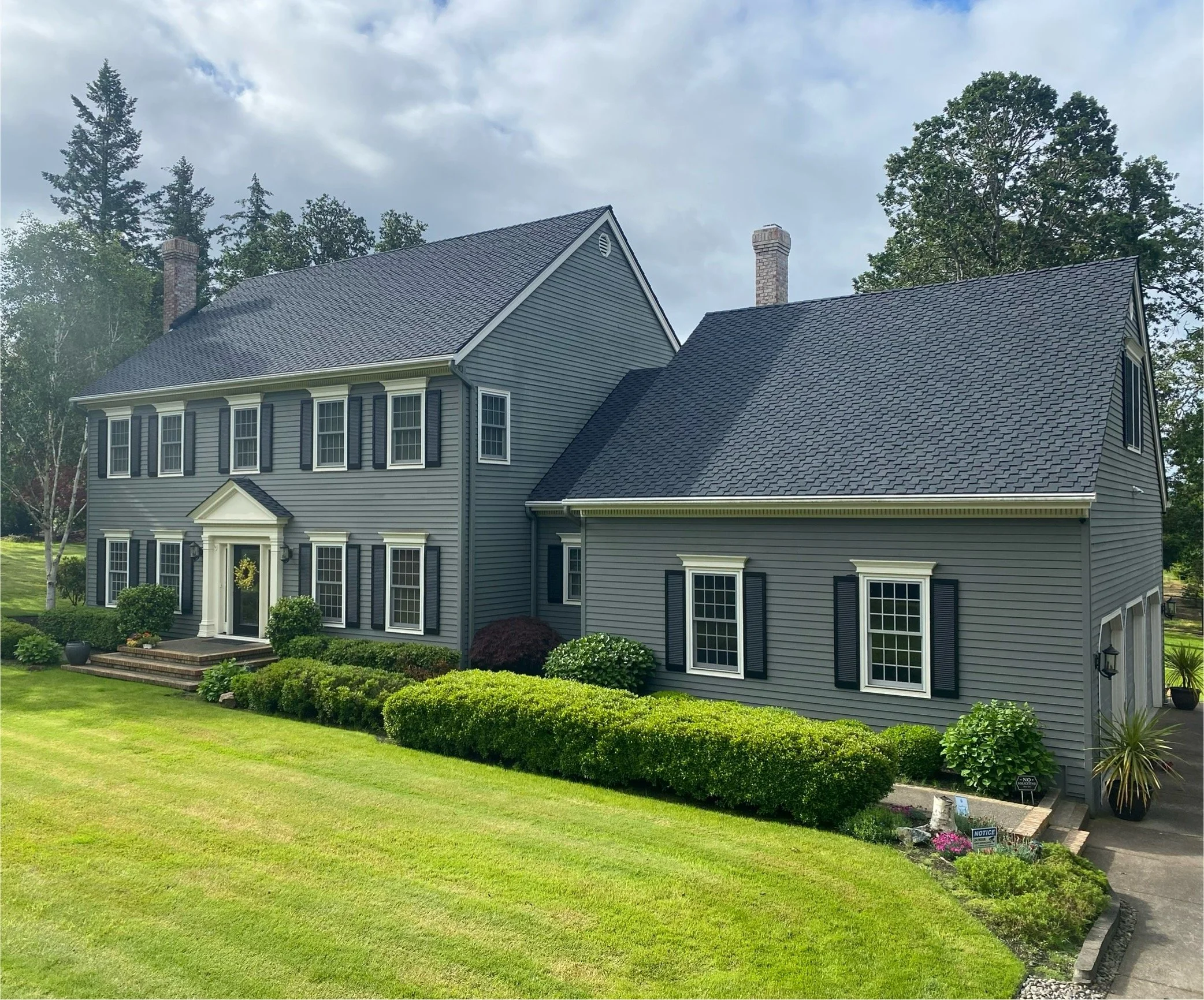 Large gray two-story house with white trim and black shutters, surrounded by a manicured lawn and shrubs, cloudy sky in the background.