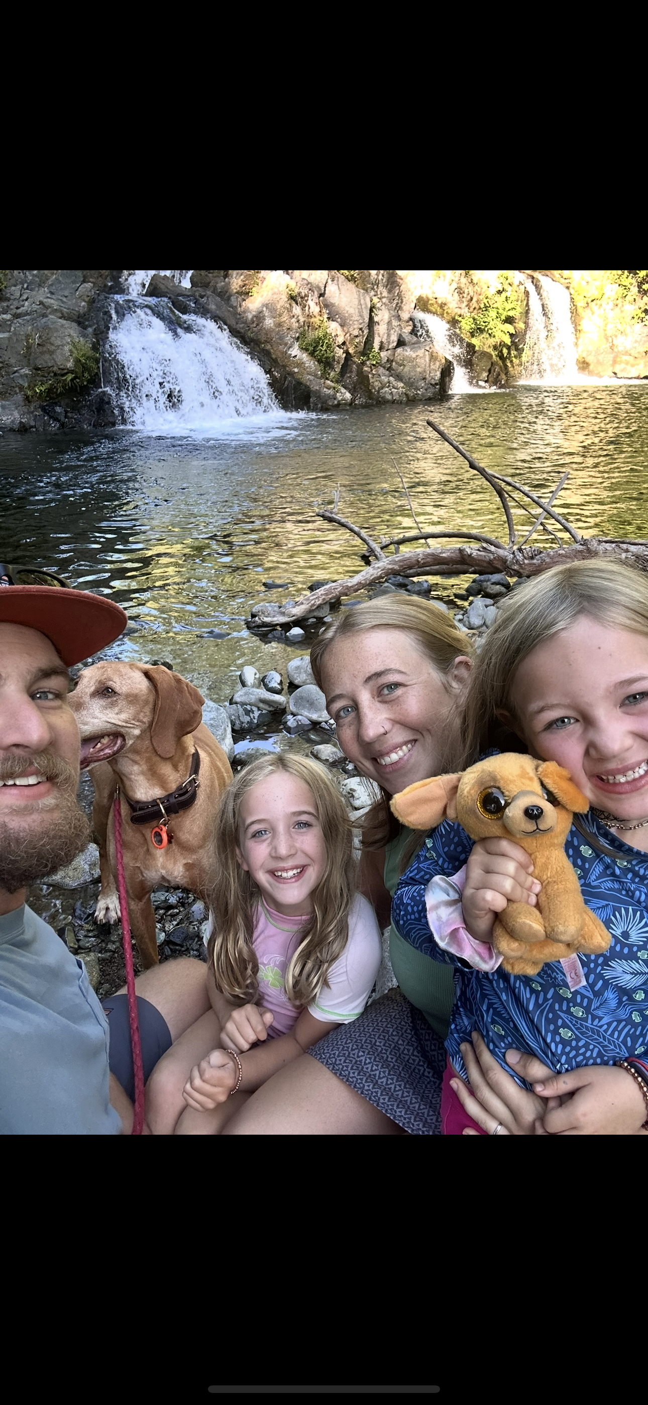 Group of five people and a dog taking a selfie in front of a waterfall by a river, with rocks and trees in the background.